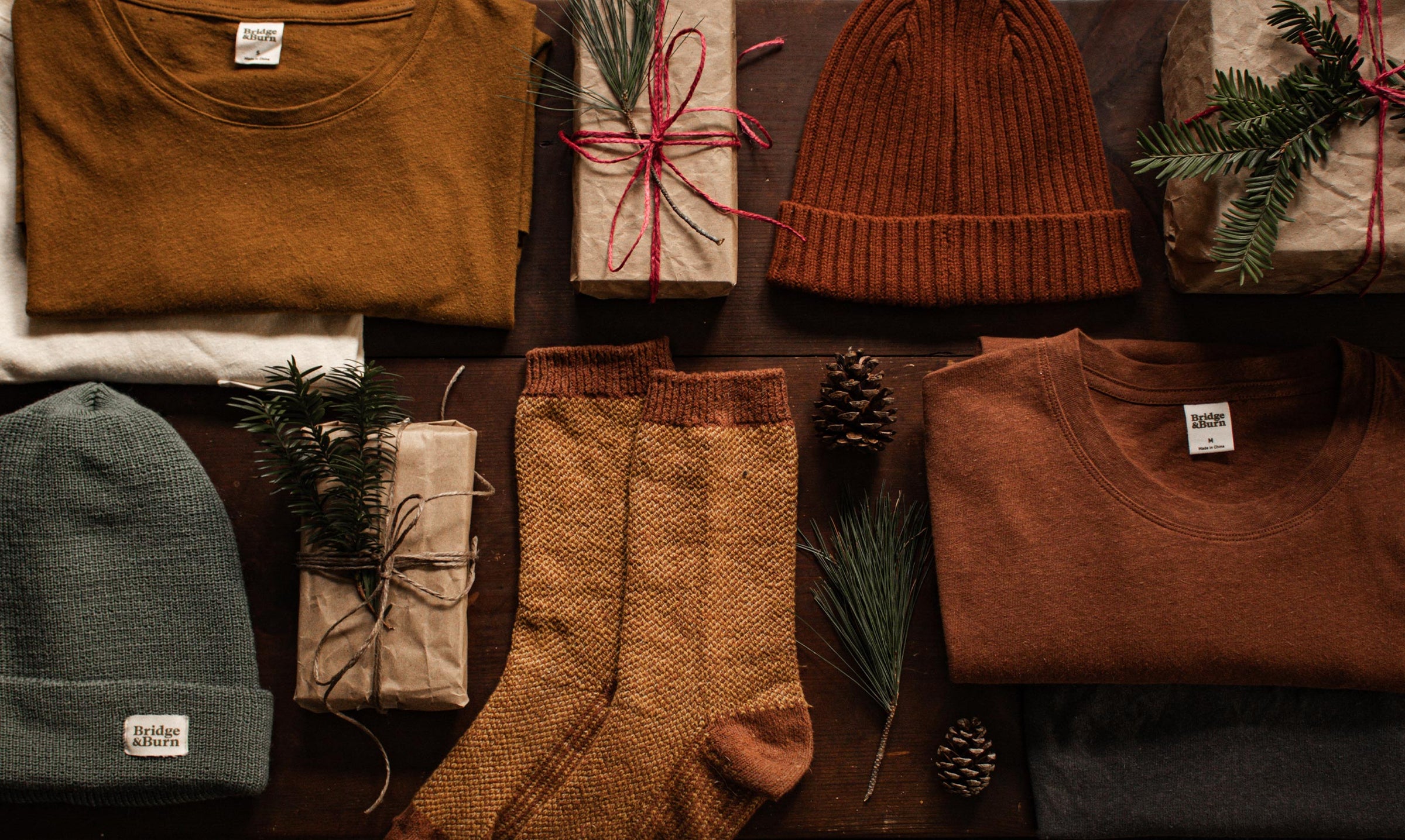 Folded shirts, knit hats, socks, and wrapped gifts with pine sprigs and pinecones are arranged on a dark wooden surface, creating a cozy, rustic holiday atmosphere.
