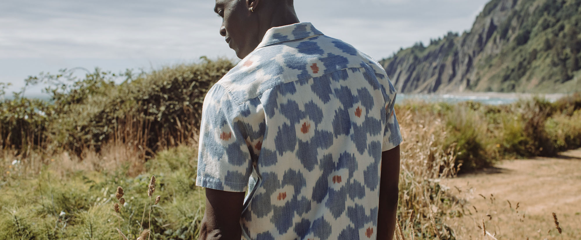 A person in a short-sleeve, patterned shirt stands outdoors facing away, with greenery, coastal cliffs, and the ocean in the background on a partly cloudy day.