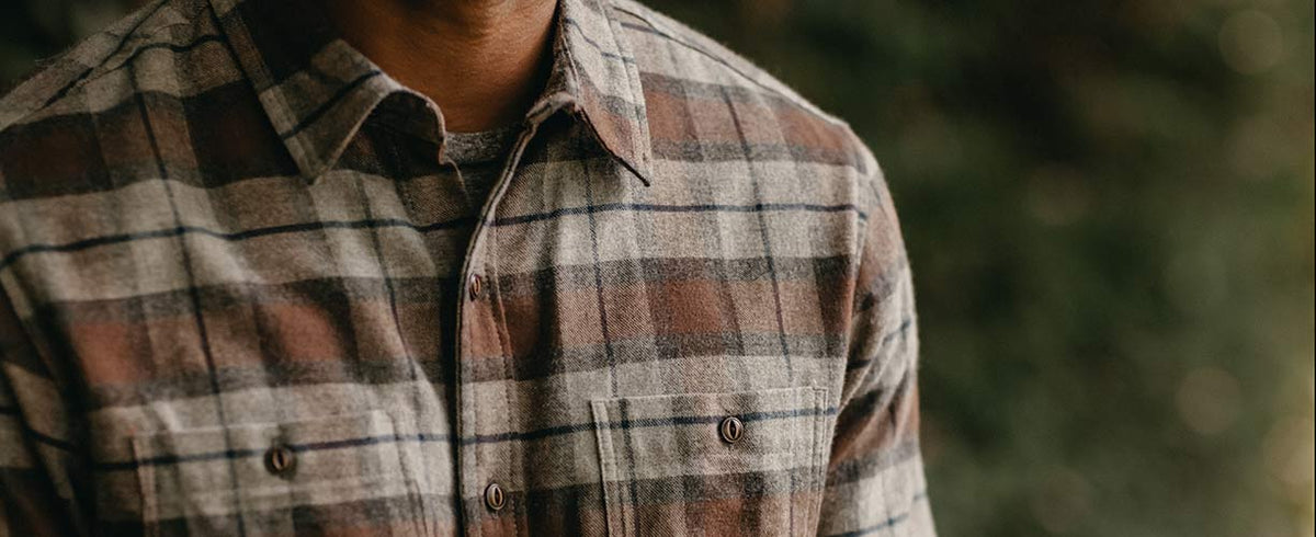 A person wearing a brown, gray, and white plaid button-up shirt stands outdoors, with the image focused on the upper torso and blurred greenery in the background.