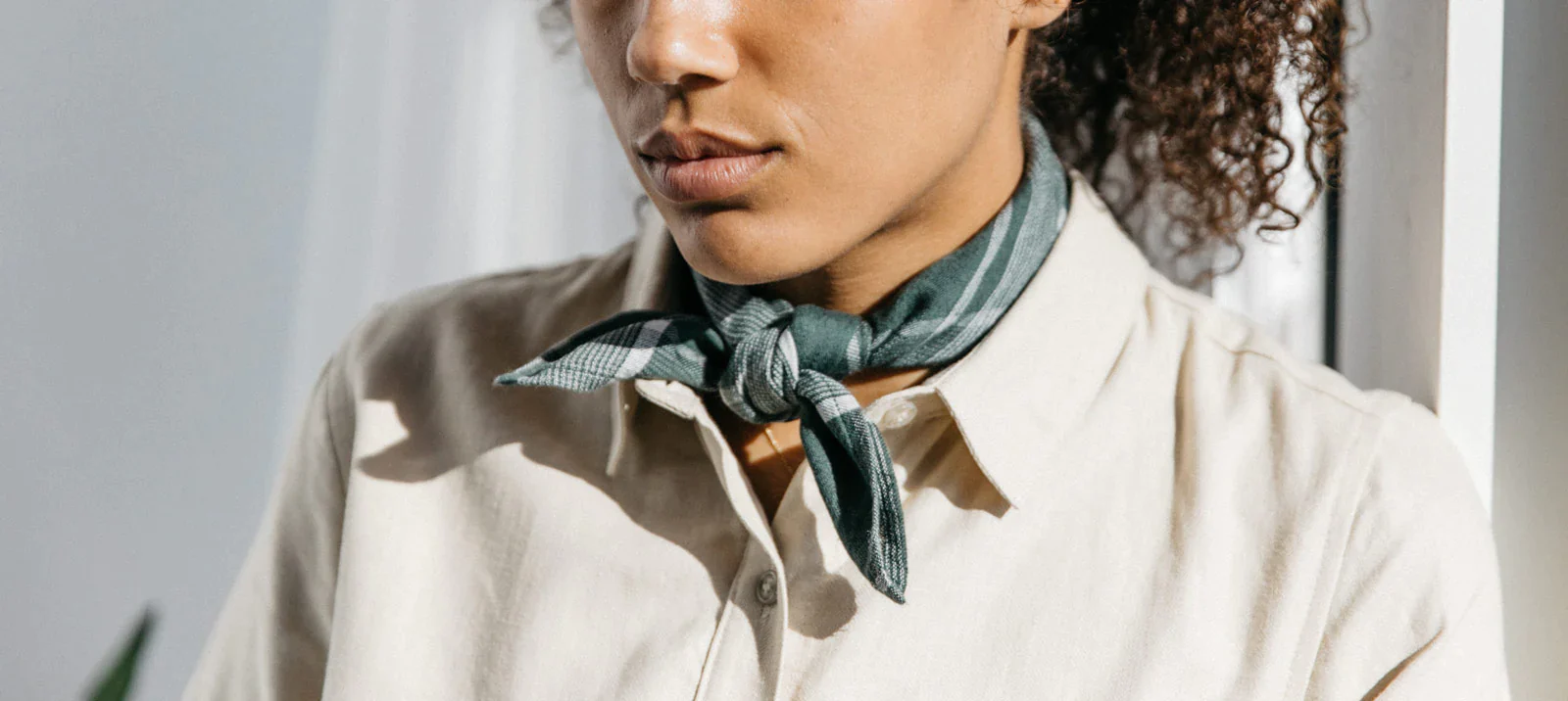 A person wearing a cream-colored button-up shirt and a green scarf tied around their neck stands near a window, with sunlight softly illuminating their face and upper body.