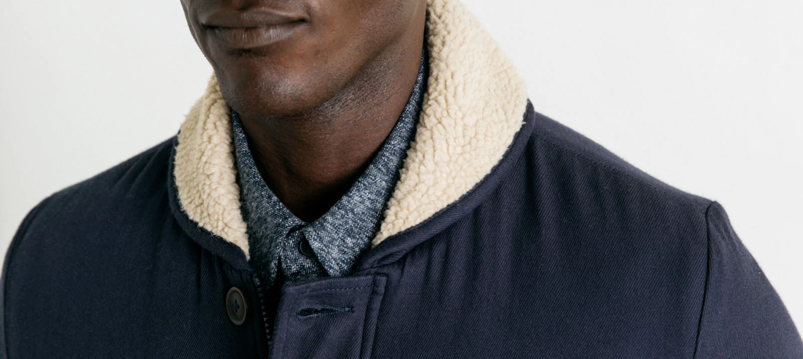 A person wearing a navy blue coat with a cream-colored sherpa collar over a blue chambray shirt, shown from the shoulders up against a plain background.