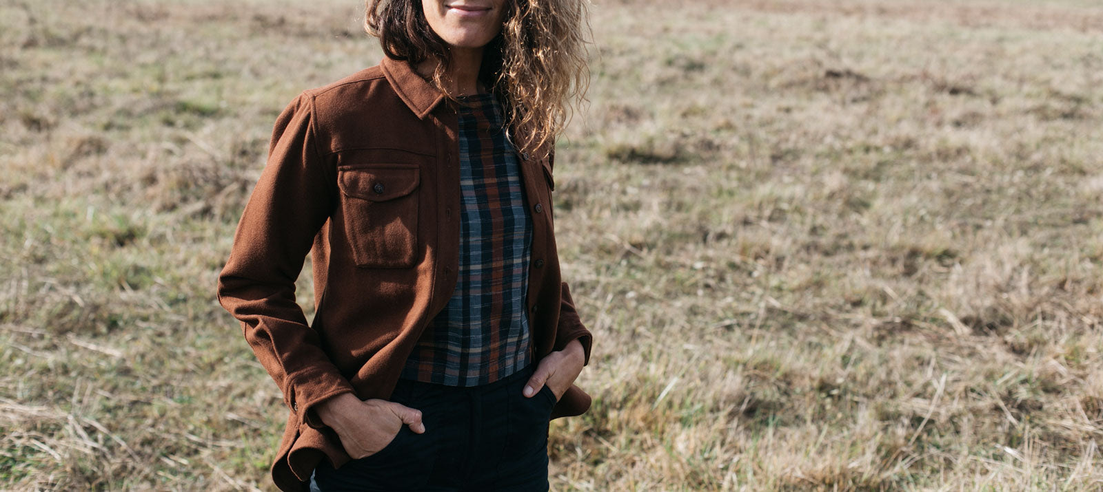 A person standing in a grassy field wearing a brown jacket over a plaid shirt with hands in their pockets. Their face is partially out of frame, and their hair is wavy.