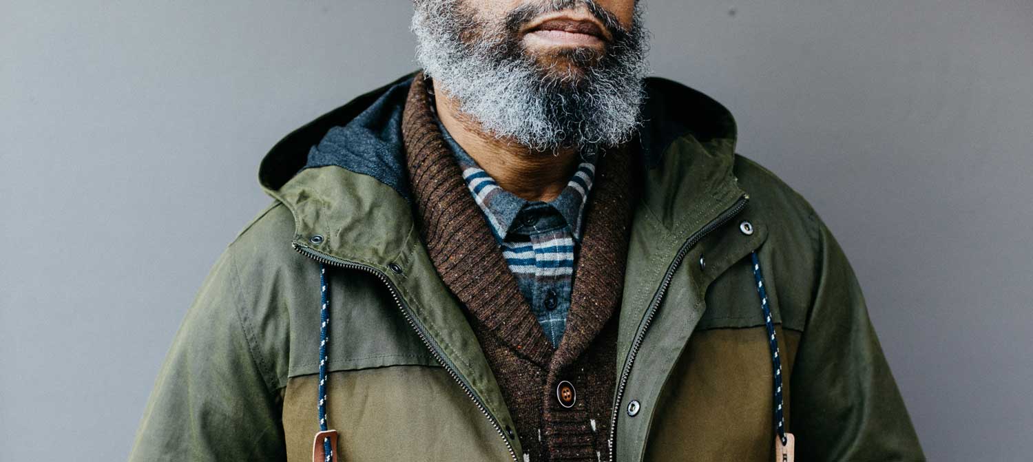 A man with a gray beard wears a green jacket over a brown sweater and a striped shirt, standing against a plain gray background. His face is partially visible from the nose down.