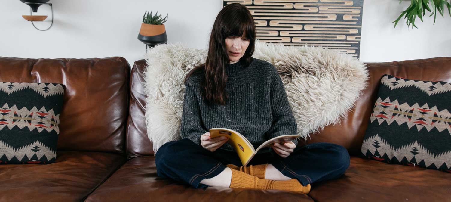 A person with long brown hair sits cross-legged on a brown leather sofa, reading a book. The sofa has patterned cushions and a fluffy throw, with plants and wall art in the background.