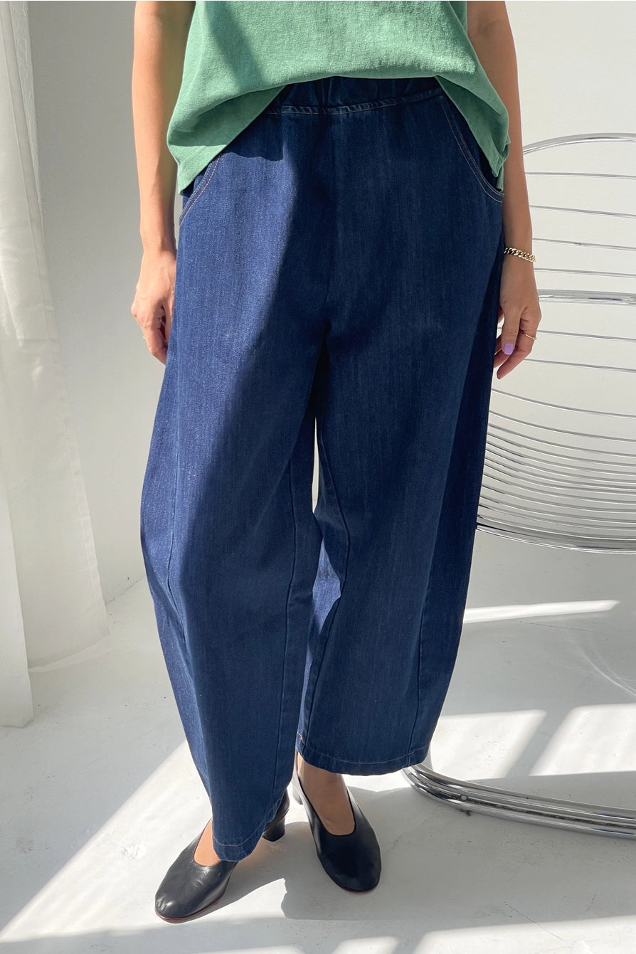 Someone stands indoors next to a metal chair, sunlight streaming onto the white floor, wearing Le Bon Shoppe - Arc Pant / Blue Denim jeans, a green top, and black shoes.