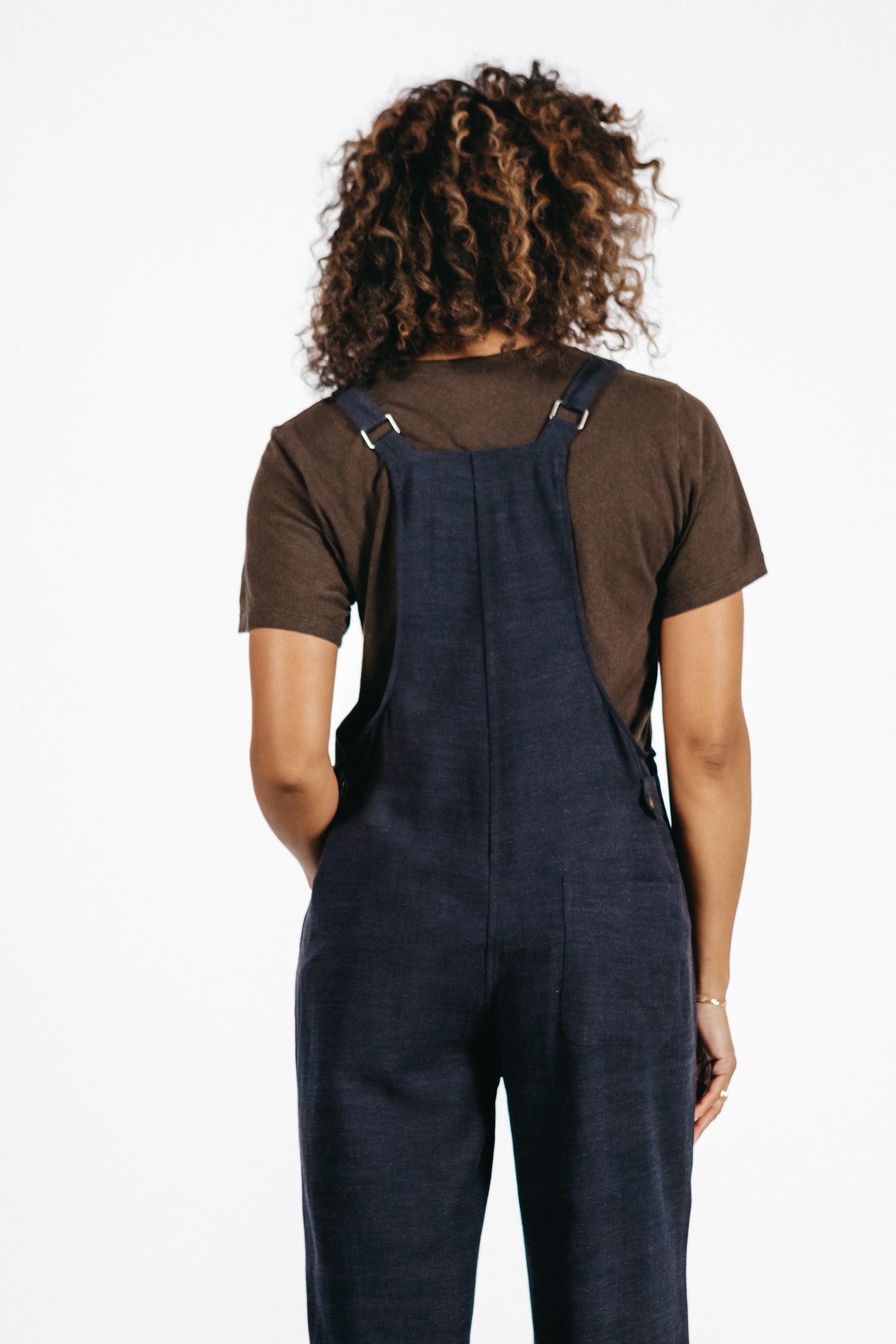 A person with curly hair, facing away, wears the Bridge & Burn Edie Overall in Navy with a brown t-shirt against a plain white background.