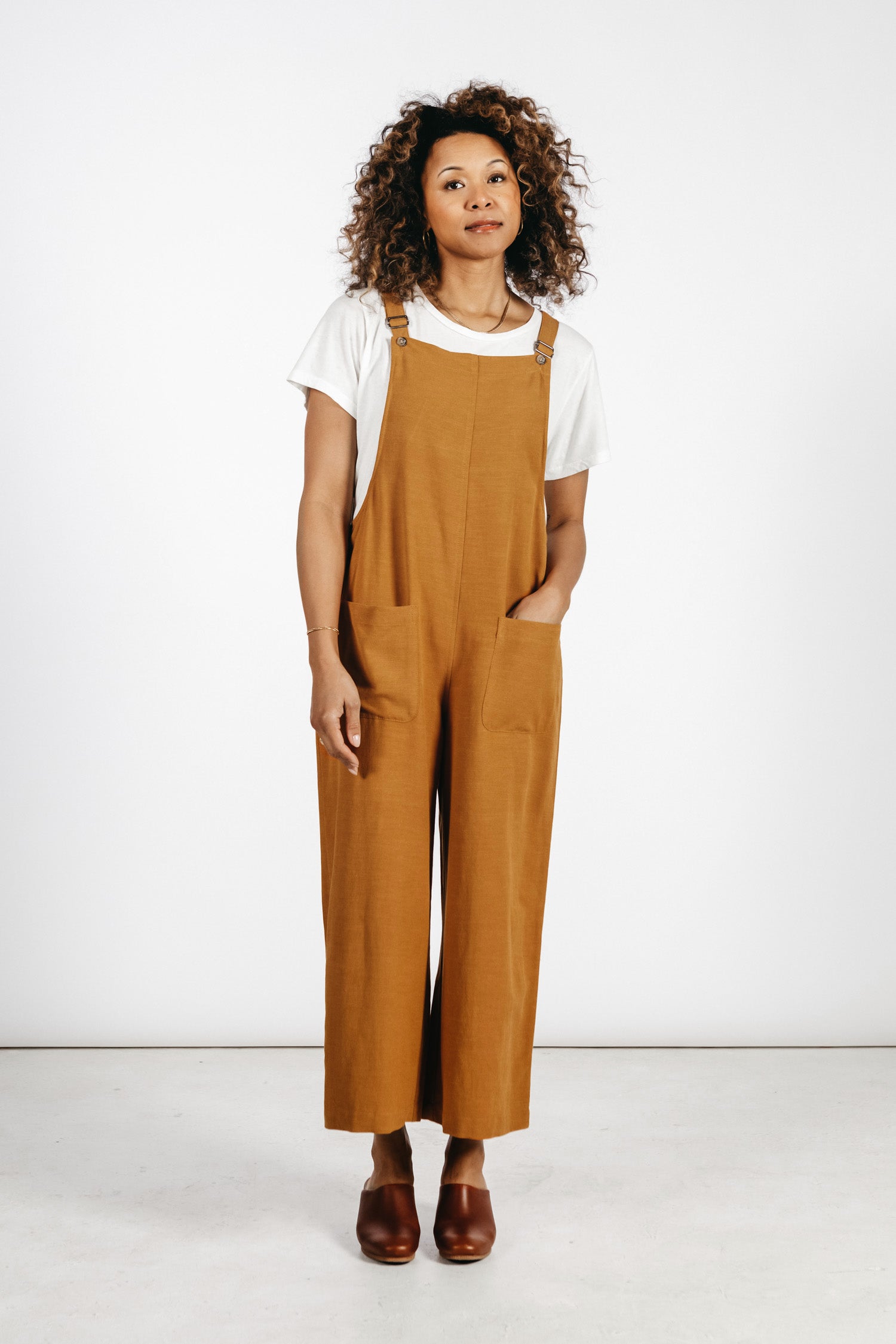 A person with curly hair stands against a white background wearing a white t-shirt, brown shoes, and the Bridge & Burn Edie Overall in Ochre, featuring wide legs, adjustable straps, and one hand in their pocket.