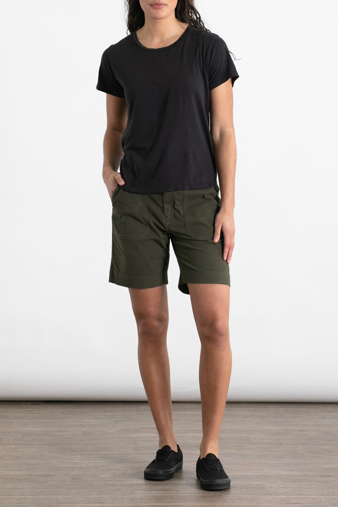 A person stands on a wood floor wearing the Jungmaven Ojai Tee in black, paired with olive green shorts and black sneakers. Their left hand is in their pocket, and their head is not visible in the image.