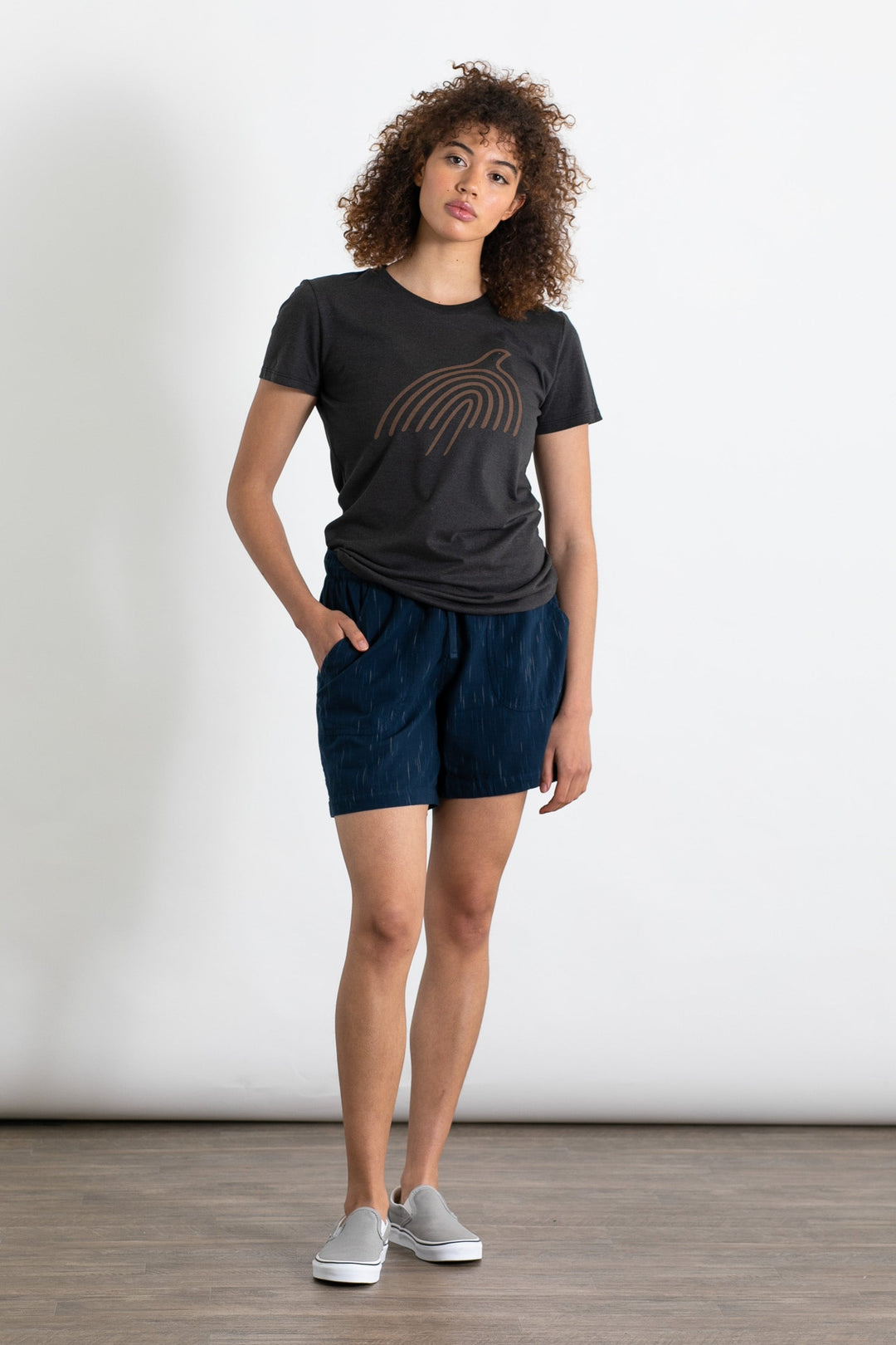 A person with curly hair wears a dark graphic tee, Bridge & Burn’s Nico Short in Navy Space Dye with an elastic waist, and gray slip-on shoes, standing against a plain white background with one hand in their pocket.
