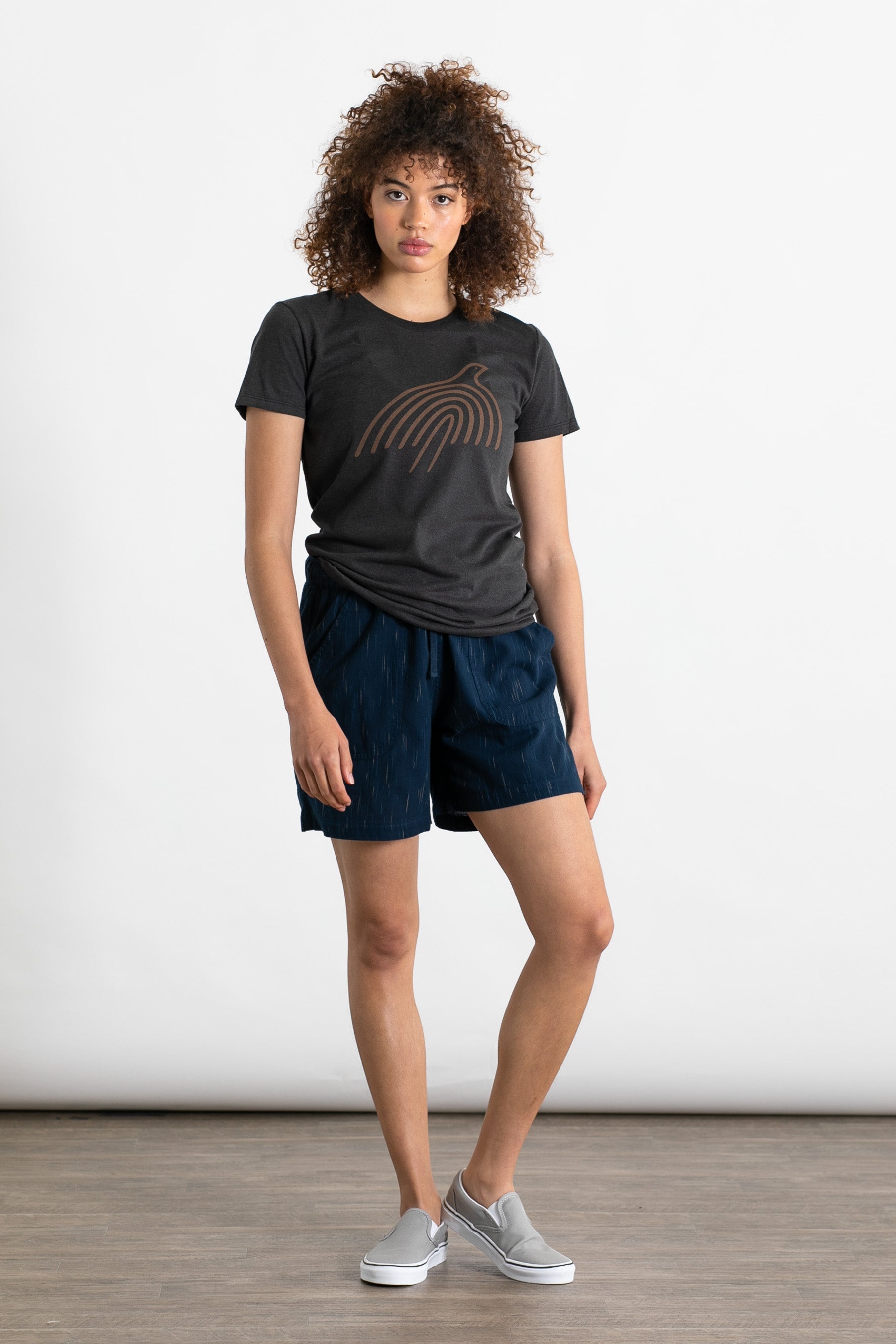 A person with curly hair wears a dark graphic tee, Bridge & Burn's Nico Short in Navy Space Dye with an elastic waist, and gray slip-on shoes, standing on a wooden floor against a plain white background.