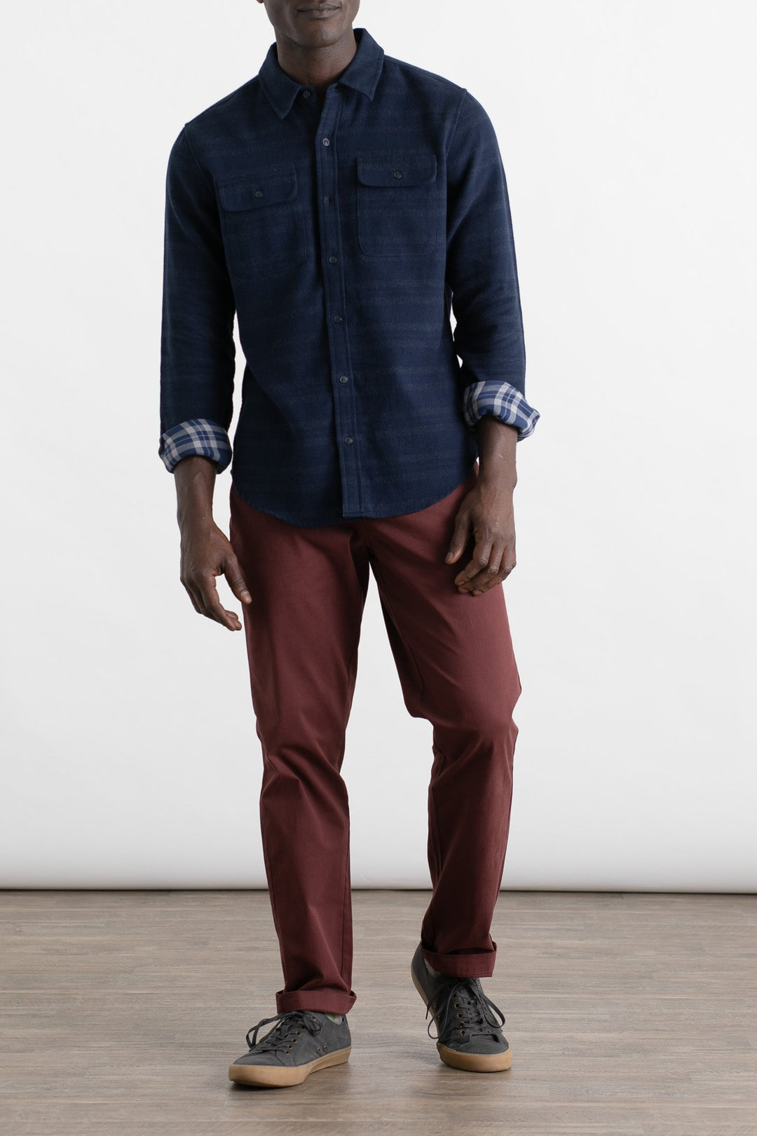 A man stands on a wooden floor against a plain light background, wearing a navy blue button-up shirt with rolled sleeves, Bridge & Burn Polk Pant in Burgundy, and black sneakers with white soles.