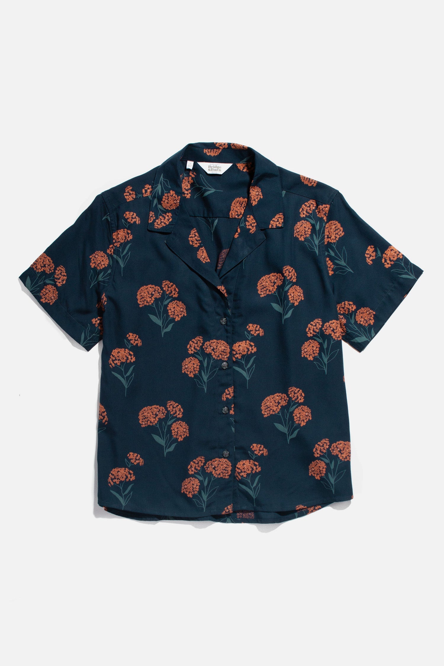 The Mila Cropped Shirt by Bridge & Burn features a relaxed fit, notched collar, and short sleeves. It’s a button-up modal shirt with a dark navy base and all-over orange and green wildflower print.