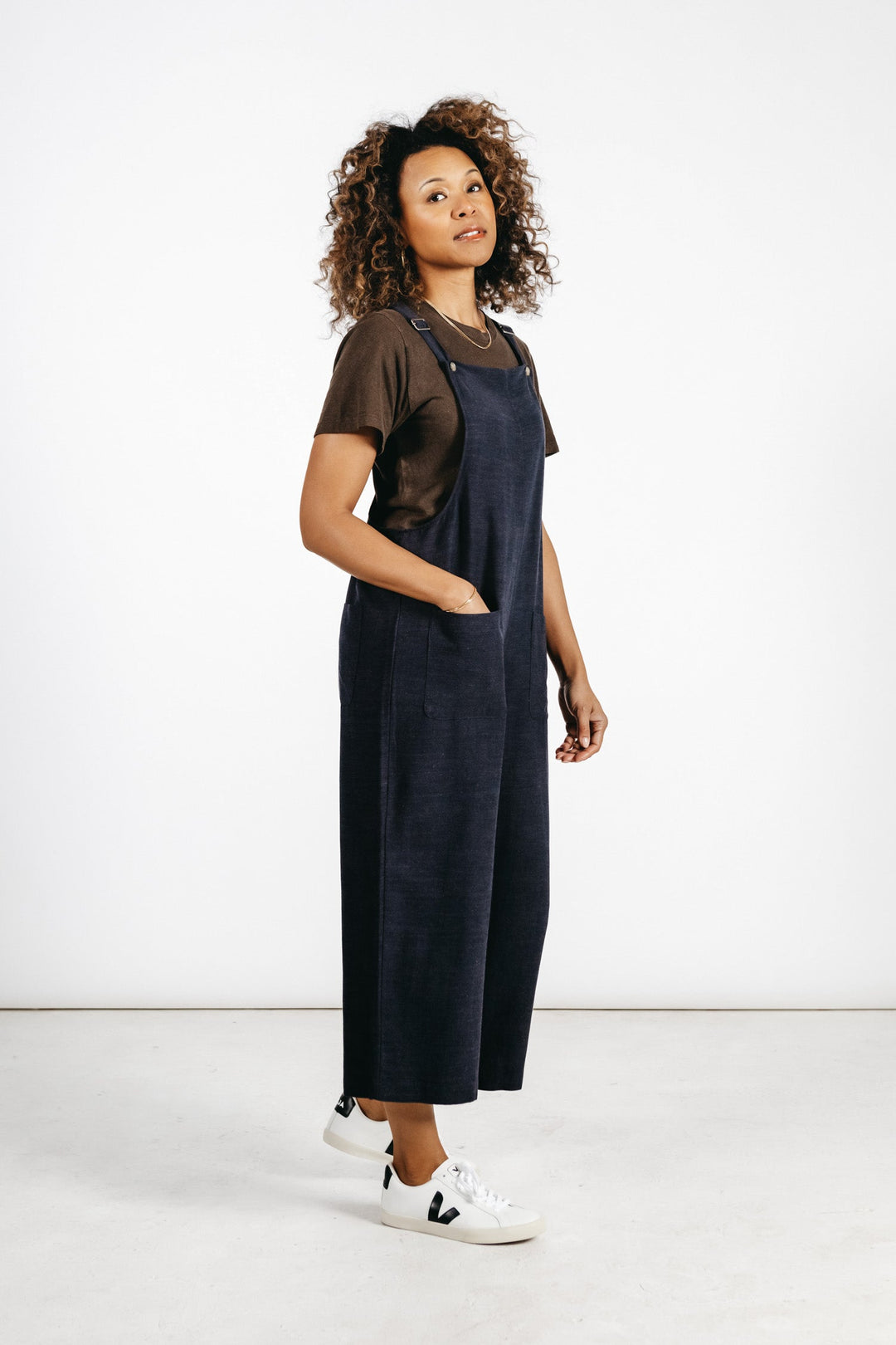 A woman with curly hair wears a brown t-shirt, white sneakers, and the Bridge & Burn Edie Overall in Navy—a wide-leg linen-cotton blend overall with adjustable straps—standing in a minimalist studio with a white background.