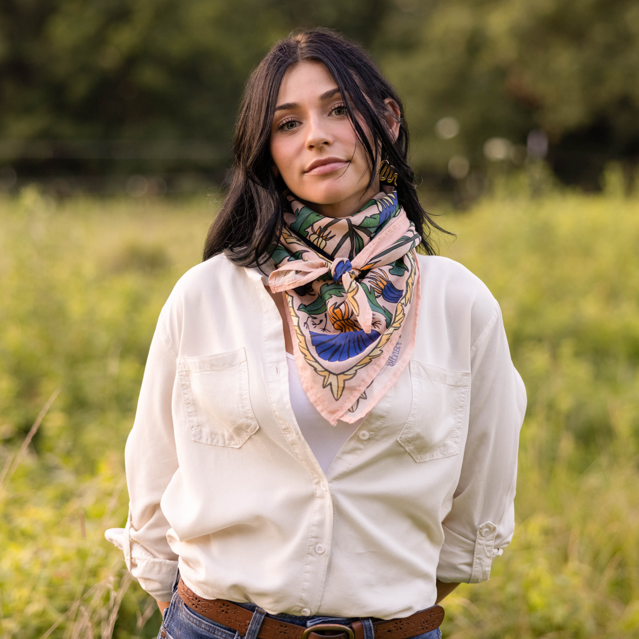 A woman with black hair stands in a grassy field, wearing a white button-up shirt and a Handker Bandanas 34" Dandy Wild Rag Bandana around her neck. She faces the camera with a neutral expression.