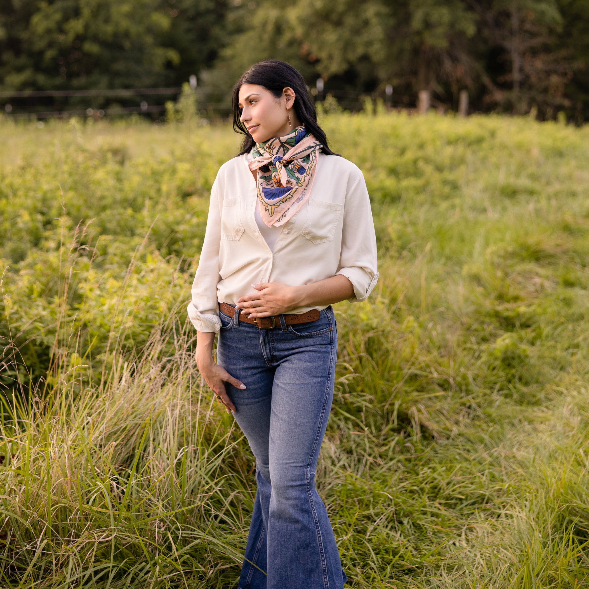 A woman stands in a grassy field, wearing a cream cotton lawn blouse, blue flared jeans, a Handker Bandanas 34" Dandy Wild Rag Bandana around her neck, and a brown belt. She gazes to the side with one hand in her pocket.