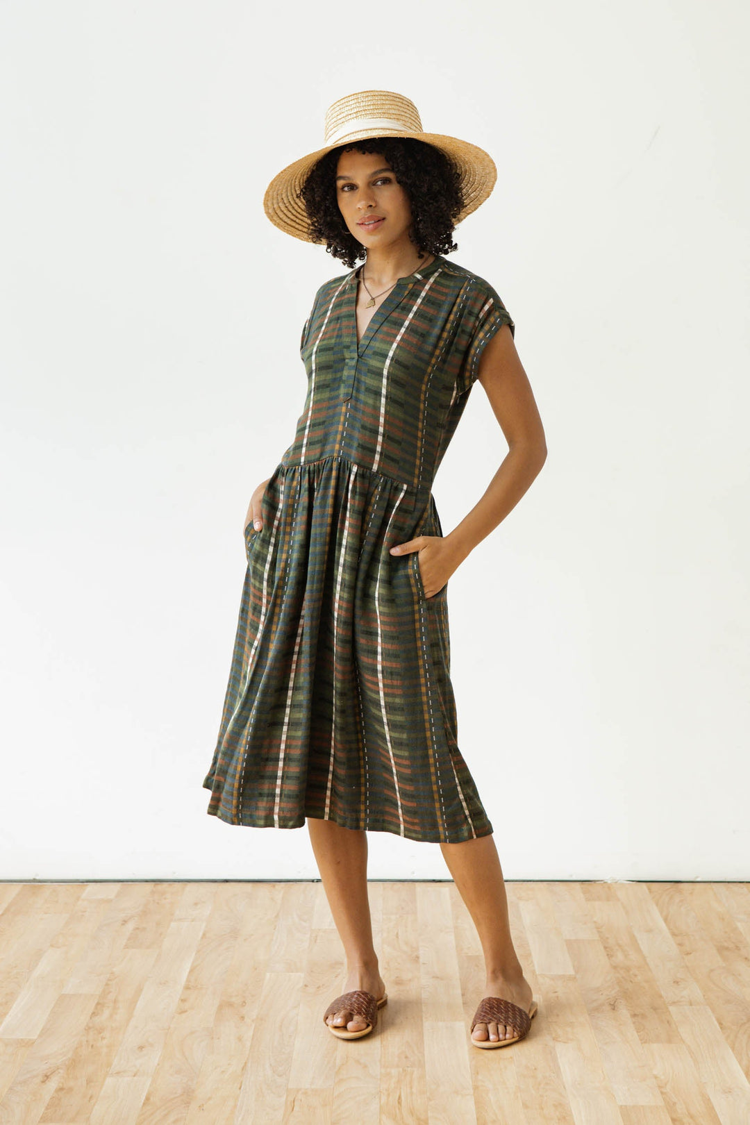 A woman in the Bridge & Burn Ira Dress in Olive Stripe—a green plaid wrinkle-resistant pullover with a gathered skirt and pockets—wears a straw hat and brown sandals, standing on a wooden floor against a plain white background.
