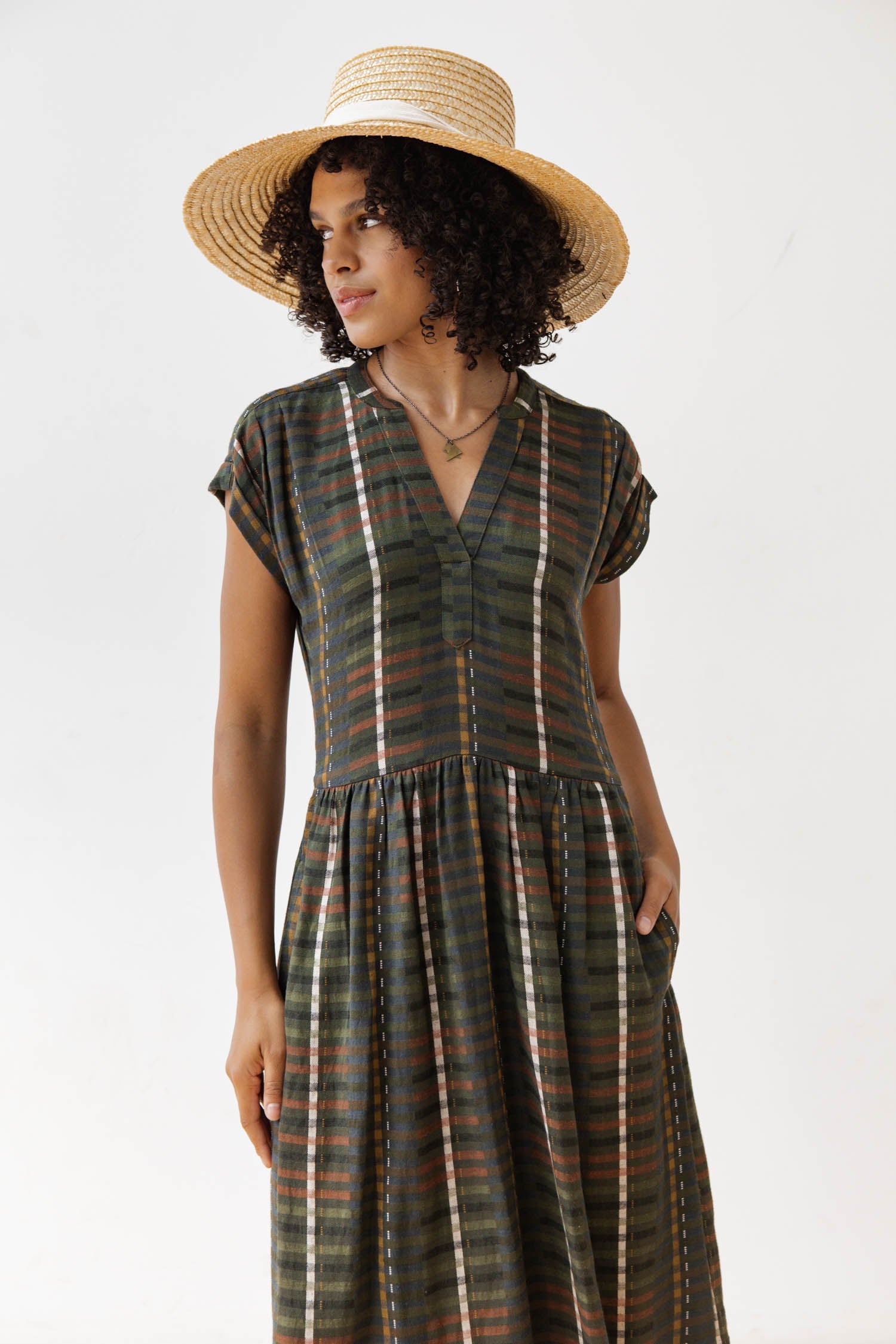 The Bridge & Burn Ira Dress in Olive Stripe is a short-sleeve, knee-length pullover with a V-neck and fitted waist, made from a modal-linen blend featuring dark green, brown, white, and rust plaid stripes.
