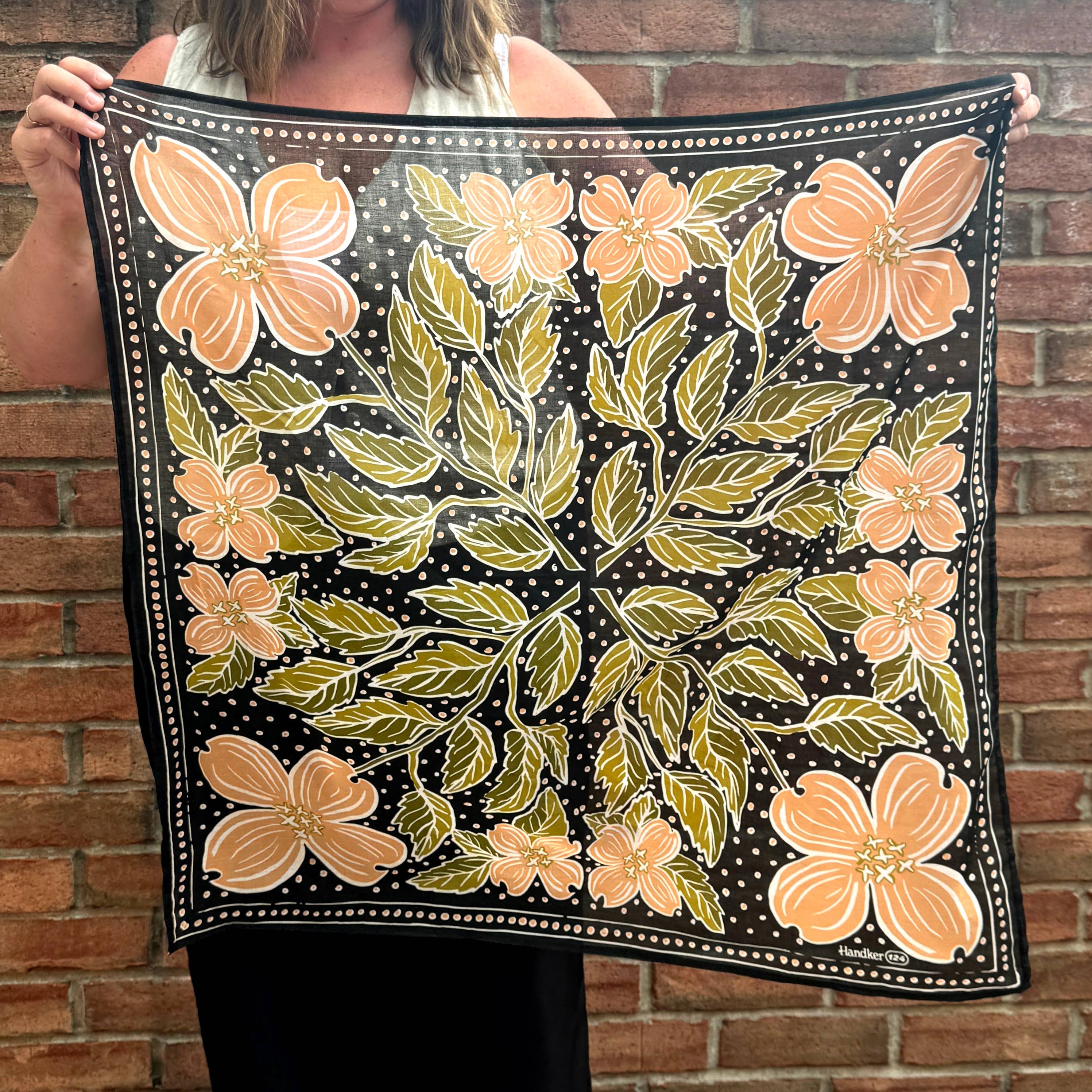 A person holds the Handker Bandanas 34" Wild Rag Amy Bandana, featuring a black background with orange flowers, green leaves, and small white dots, while standing in front of a brick wall.