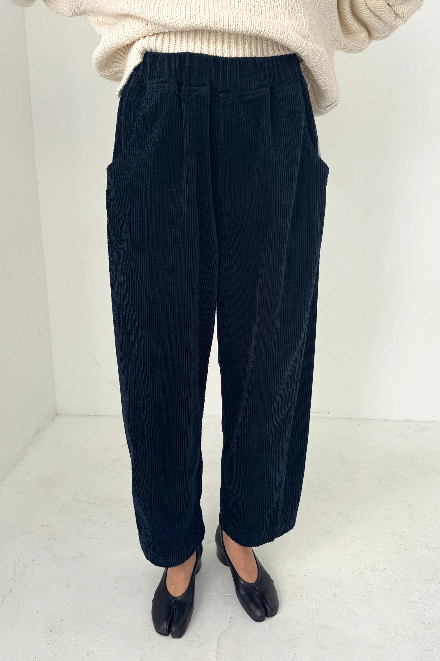 Someone wearing Le Bon Shoppe’s Arc Pant in Midnight Corduroy, a cream knit sweater, and black split-toe shoes stands in a minimalist, white-walled room.