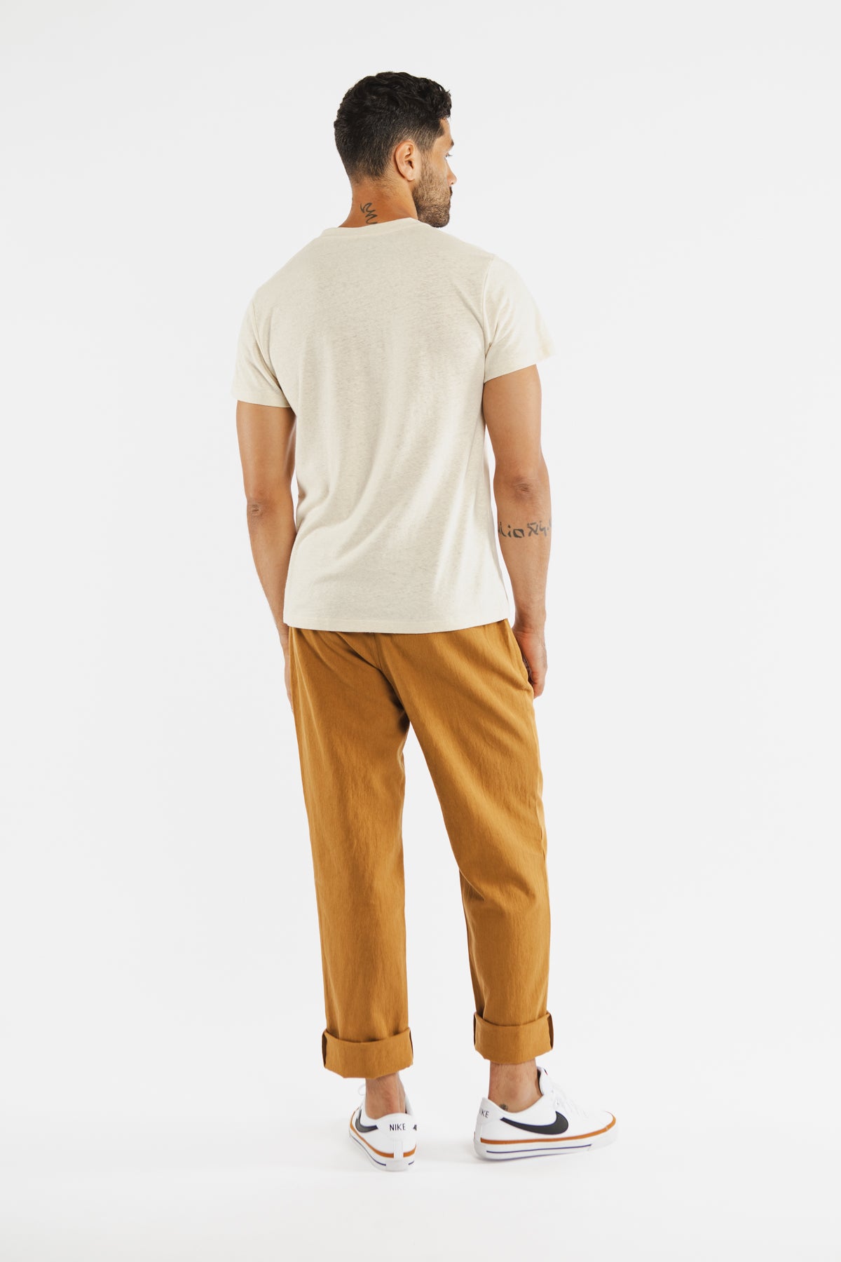 A man faces away from the camera, wearing the Bridge & Burn Organic Hemp Pocket Tee in Ivory. He pairs it with rolled-up brown pants and white Nike sneakers with navy swooshes. The background is plain white.