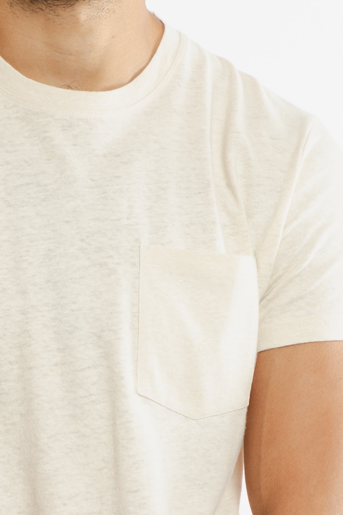 A close-up of a person wearing the Bridge & Burn Organic Hemp Pocket Tee in Ivory, featuring a small chest pocket, with part of their neck, chest, and upper arm visible against a plain background.