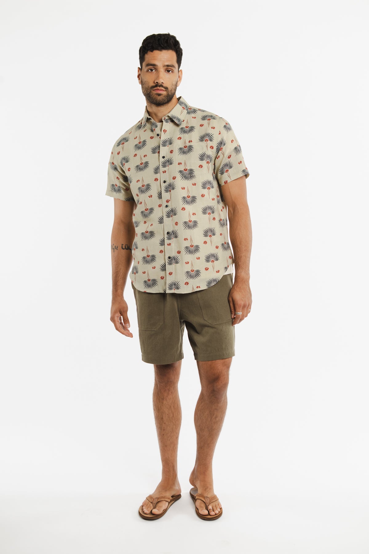 A man with a short beard and curly hair stands against a white background, wearing the Bridge & Burn Harbor Slim Shirt in Aloha Floral. The shirt is slim fit with short sleeves, paired with olive green shorts and brown flip-flops.