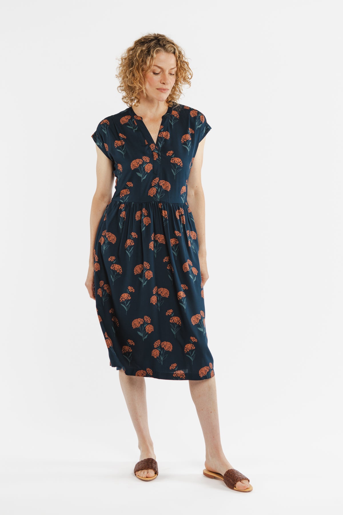 The Bridge & Burn Ira Dress / Wildflower is a navy blue knee-length modal pullover with short sleeves, a V-neckline, an elastic waist, and an orange and green floral print. Shown laid flat on a white background.