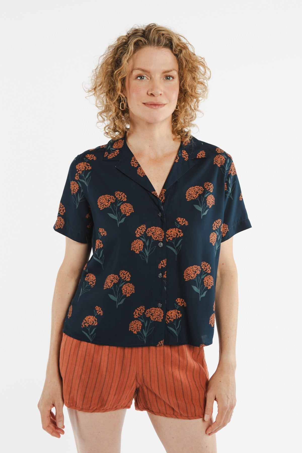 A person with curly blonde hair wears the Bridge & Burn Mila Cropped Shirt in the Wildflower pattern and rust-colored shorts, standing against a plain white background.