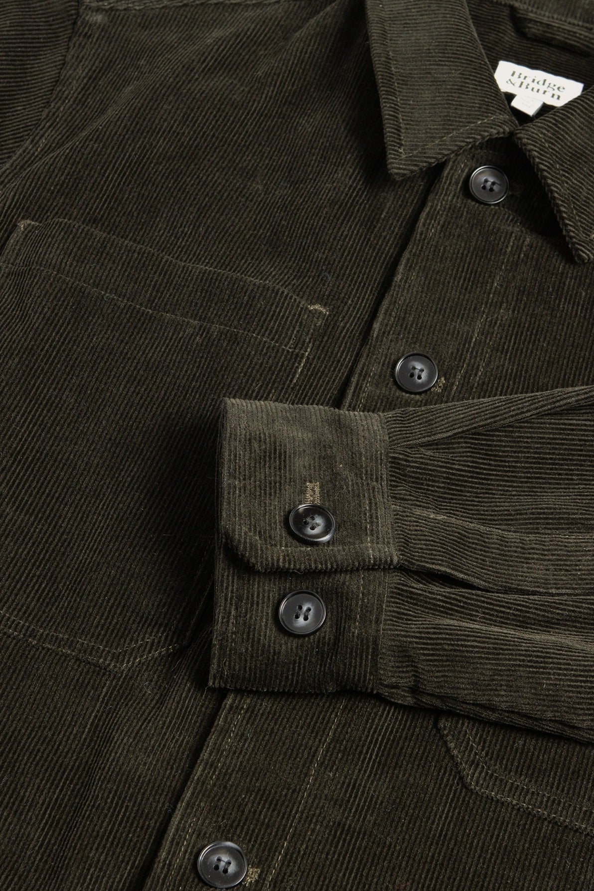 A close-up of the Bridge & Burn Banks Shirt Jacket in Deep Pine Corduroy shows its black buttons, chest pocket, and ribbed texture. One sleeve is folded across the body for a workwear look. The garment label is partially visible at the collar.