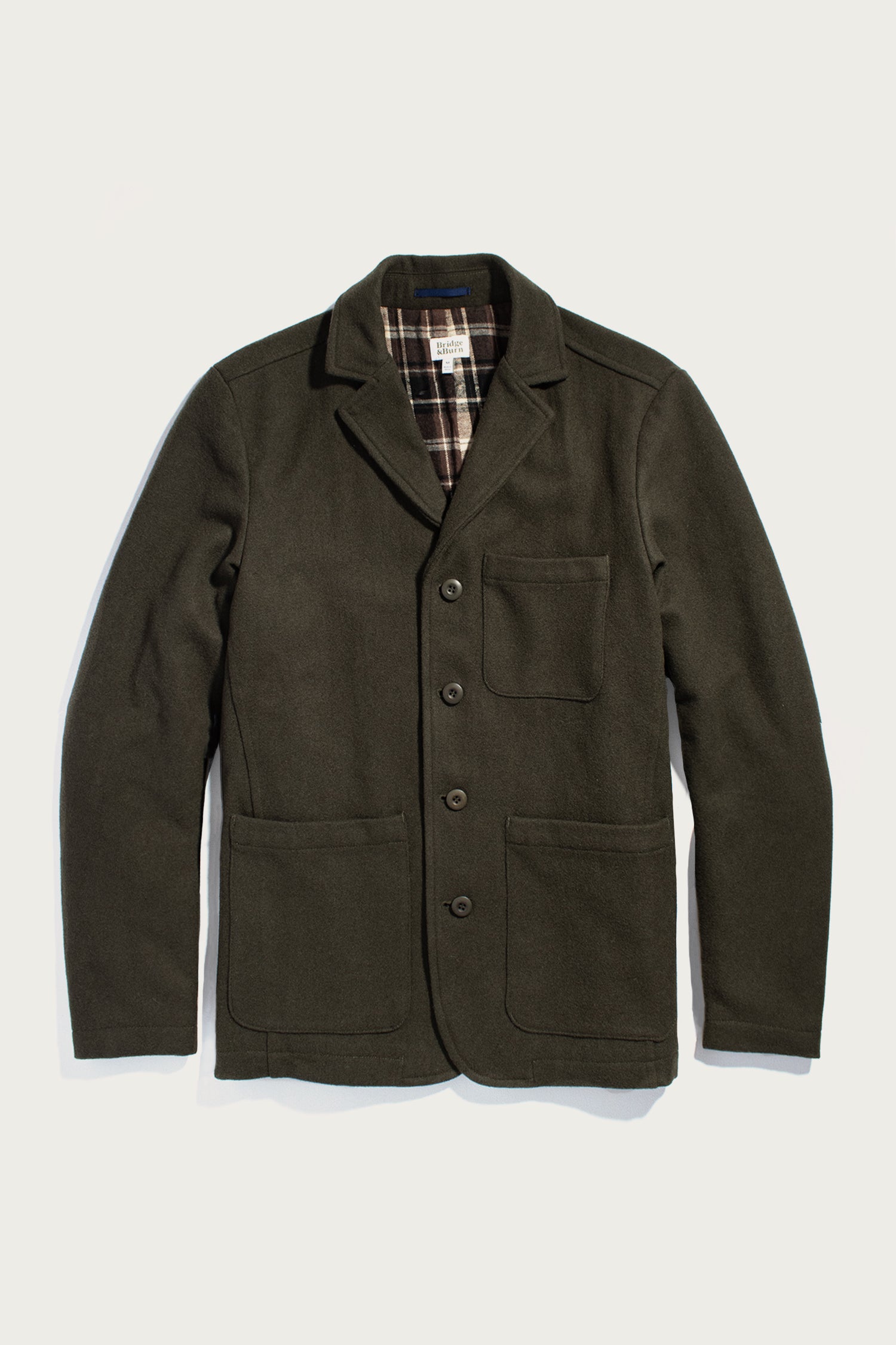 The Bridge & Burn Nate Chore Blazer in Basque Green wool features a dark green hue, three front pockets, button closure, and a plaid lining. Shown flat on a white background.
