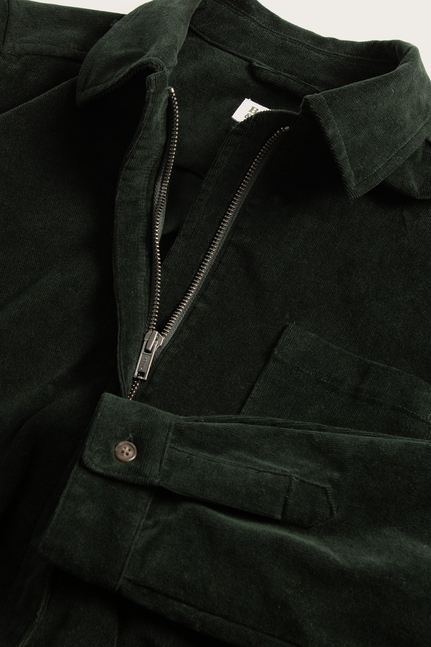 A close-up of the Bridge & Burn Faye Jumpsuit in Deep Pine Pinwale, a dark green stretch corduroy style with a half-zip front, chest pocket, and buttoned sleeve cuffs.