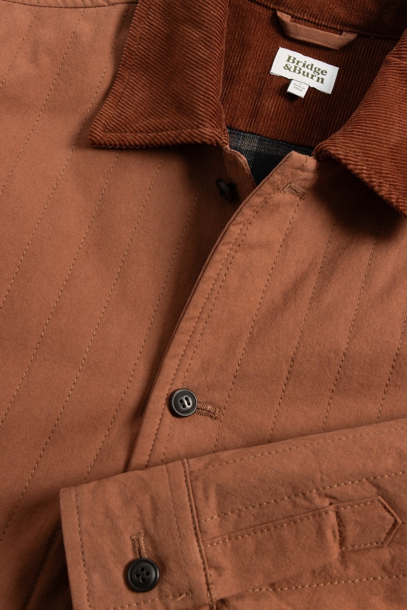 Close-up of the Kenton Jacket in Sienna—a brown quilted jacket with a corduroy collar, black buttons, and a white label inside the collar showing the Bridge & Burn brand. This lightweight piece offers both style and comfort.