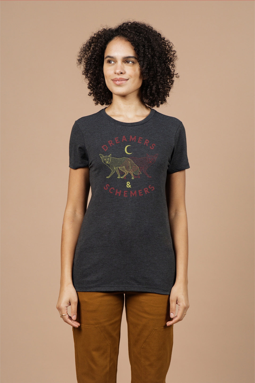A woman with curly hair wears the Bridge & Burn Women's Dreamers & Schemers Tee in Black, featuring a fox, crescent moon, and red-yellow "DREAMERS & SCHEMERS" graphic. She stands against a beige background, looking slightly to the side.