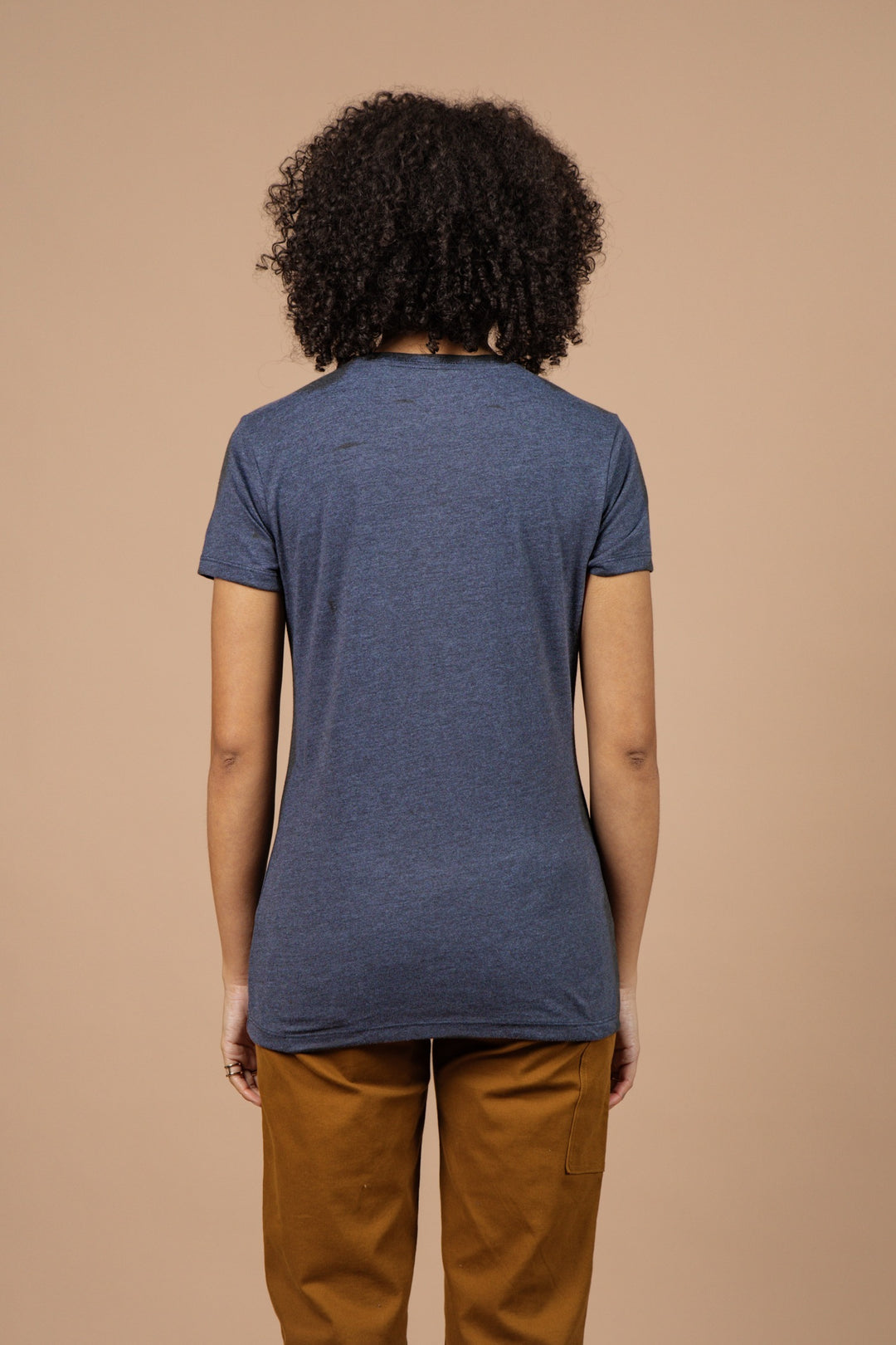 A woman with curly hair, wearing Bridge & Burn’s Women's Tallest Tree Tee in navy, stands with her back to the camera against a plain beige background, paired with brown pants.