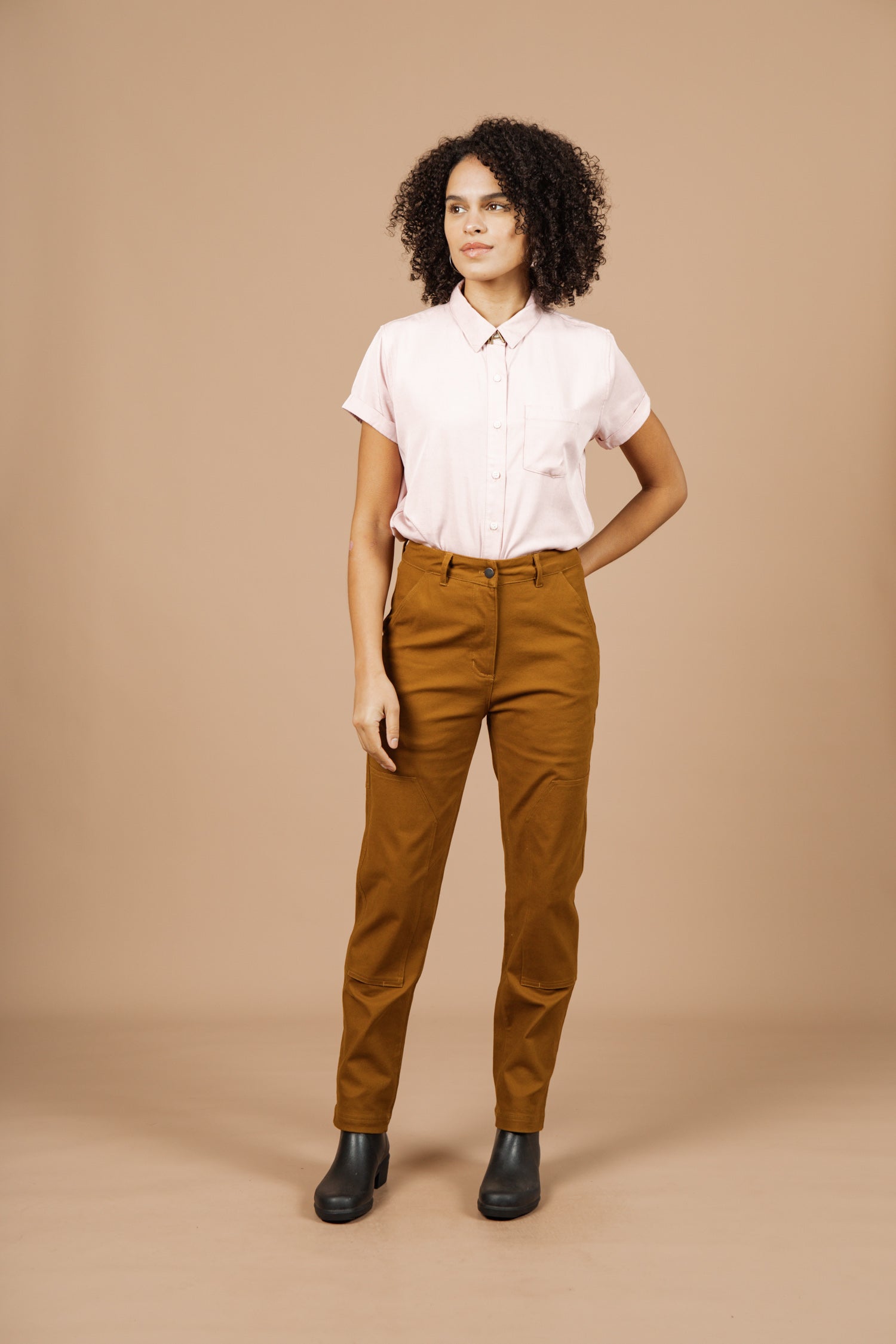 A person with curly hair stands against a beige background, wearing a light pink short-sleeved button-up shirt and Bridge & Burn Bowden Utility Pant in Hickory Canvas with reinforced knees, paired with black ankle boots and one hand in their pocket.