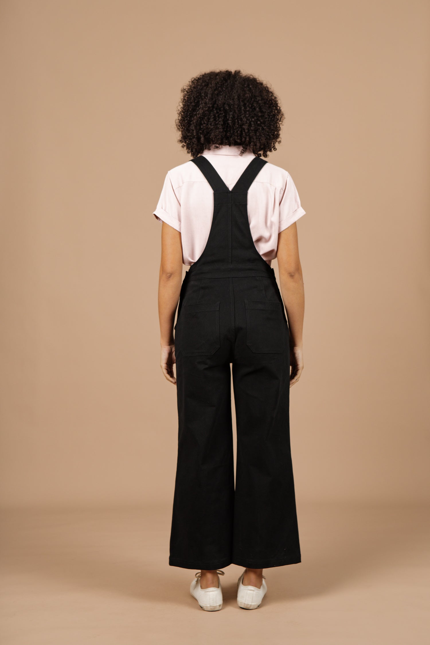 A person with curly hair, facing away, wears a light pink short-sleeve shirt and Bridge & Burn's Roscoe Overall in Black Canvas paired with white shoes, standing against a plain beige background.