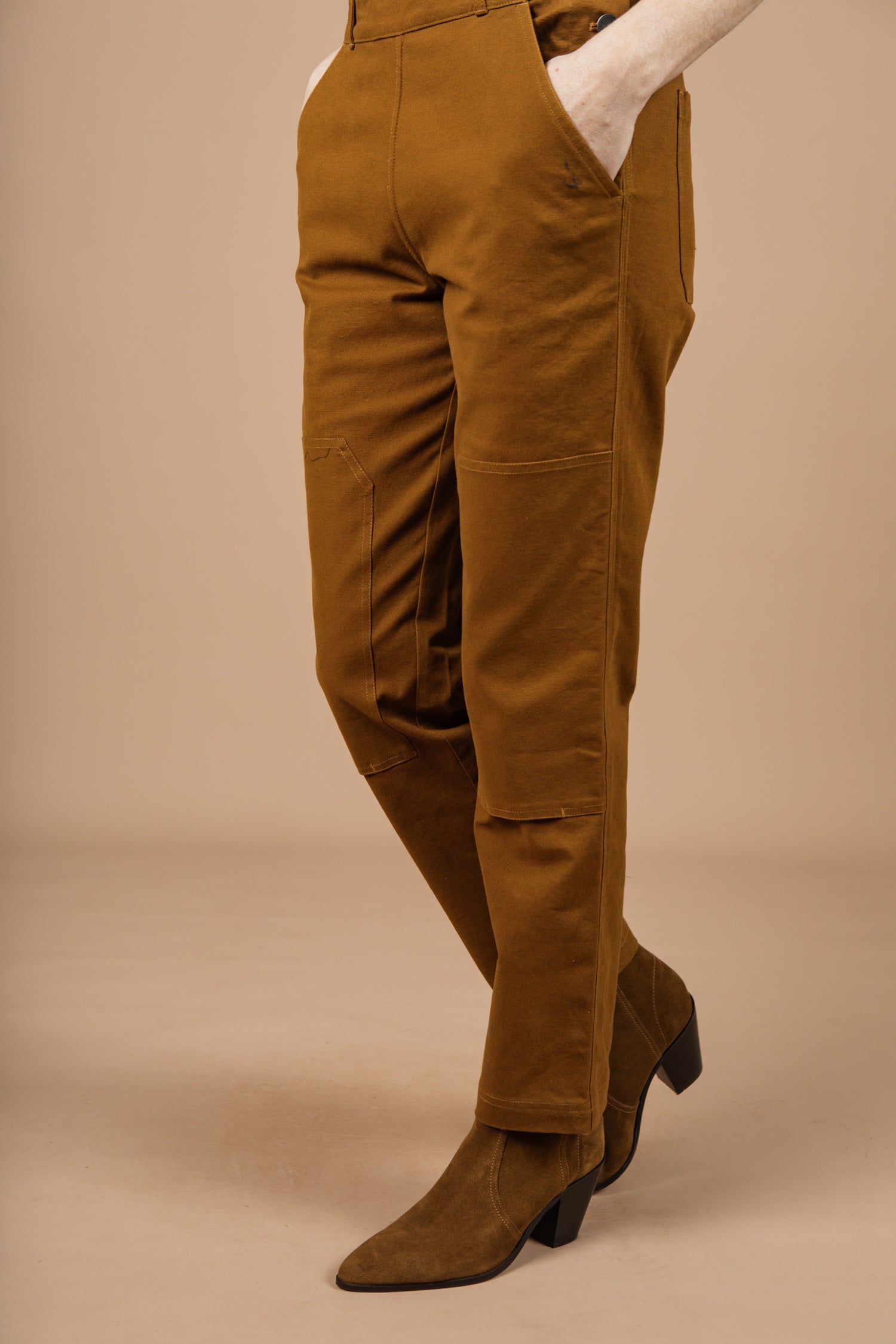 A person wears the Bridge & Burn Tobin Utility Overall in brown canvas—high-waisted with visible front seams and pockets—paired with brown suede heeled boots, standing against a beige background.