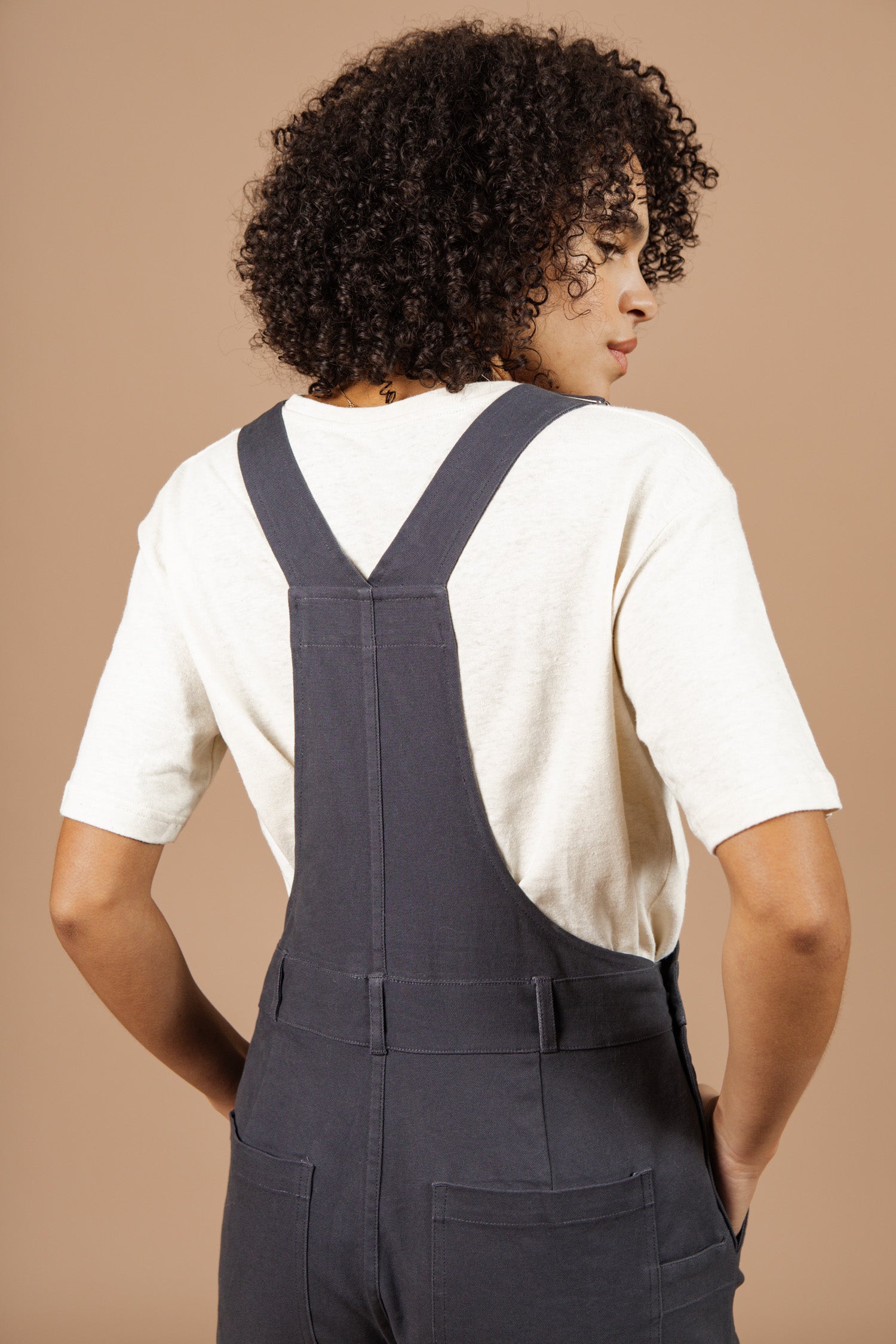 A person with curly hair, shown from the back against a beige background, wears a cream short-sleeve shirt and the Bridge & Burn Tobin Utility Overall in Dark Charcoal Canvas.