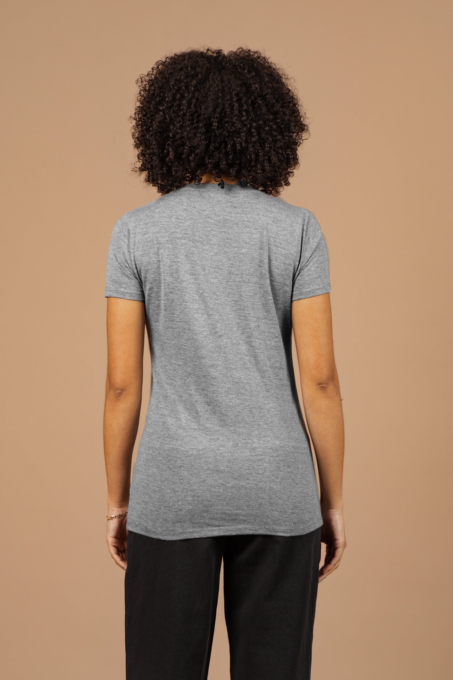 A person with curly hair, wearing the Bridge & Burn Women's Grown Tee in grey and black pants, stands facing away from the camera against a plain beige background.