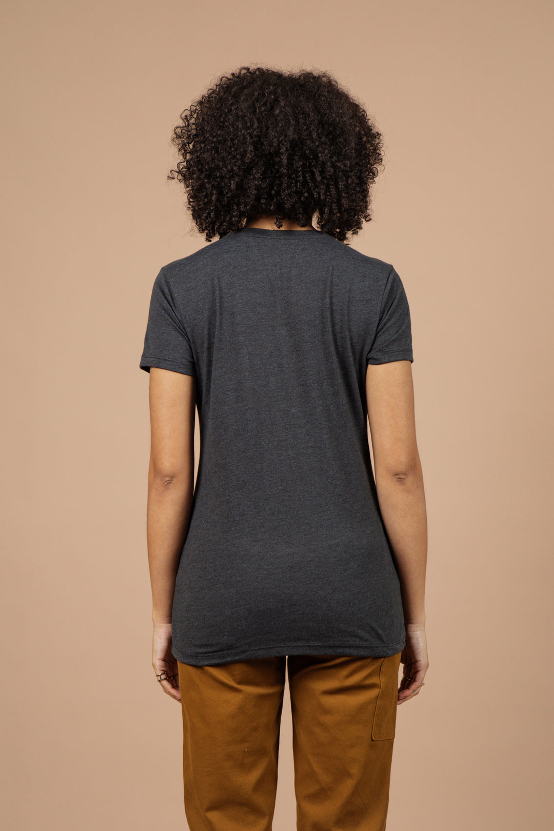 A person with curly hair stands with their back to the camera against a plain beige background, wearing the Bridge & Burn Women's Mycology Tee in black and brown pants.