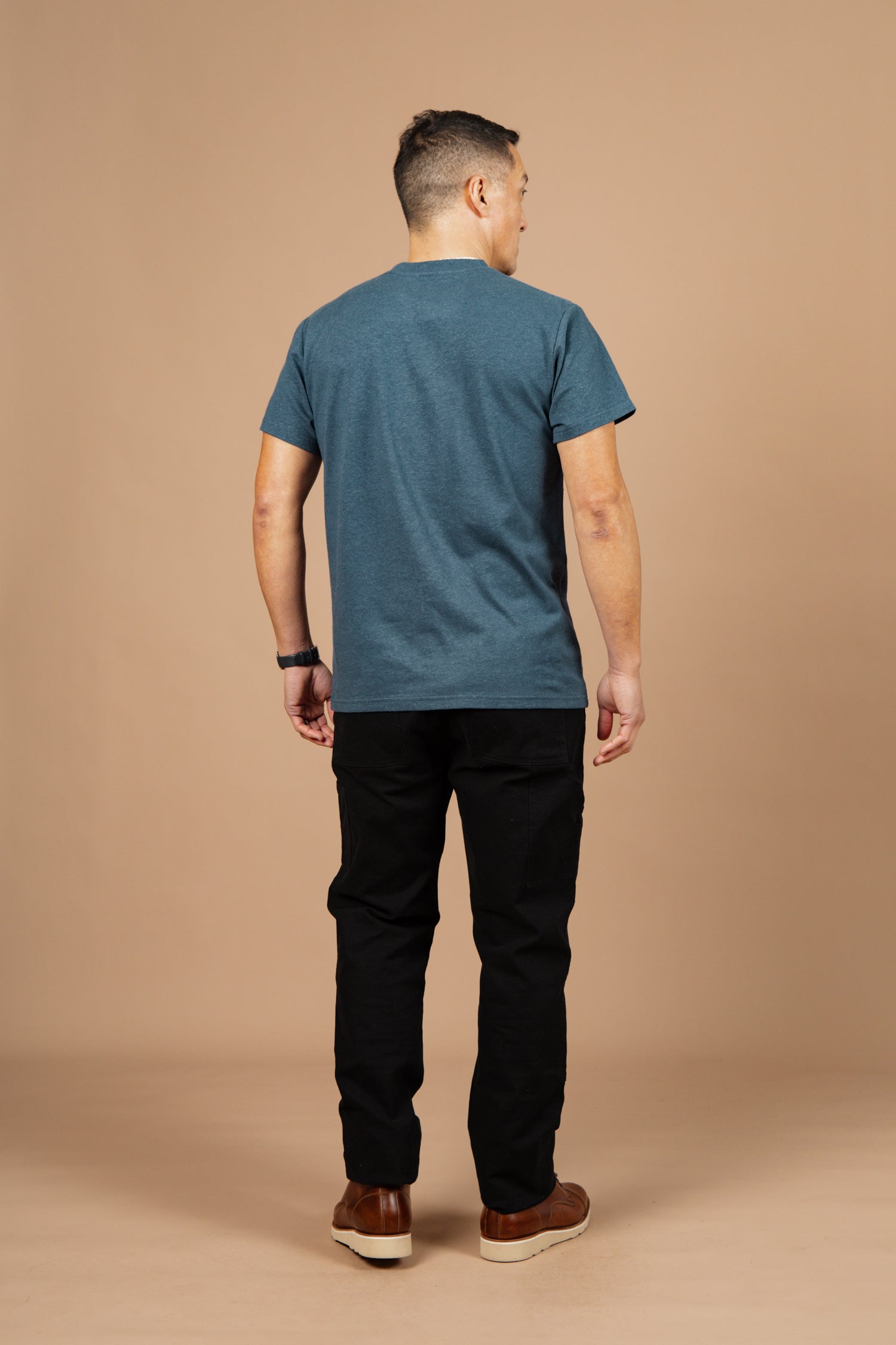 A man stands facing away from the camera, wearing the Bridge & Burn Organic Hemp Pocket Tee in Storm Blue, black pants, brown shoes, and a watch against a plain beige background.