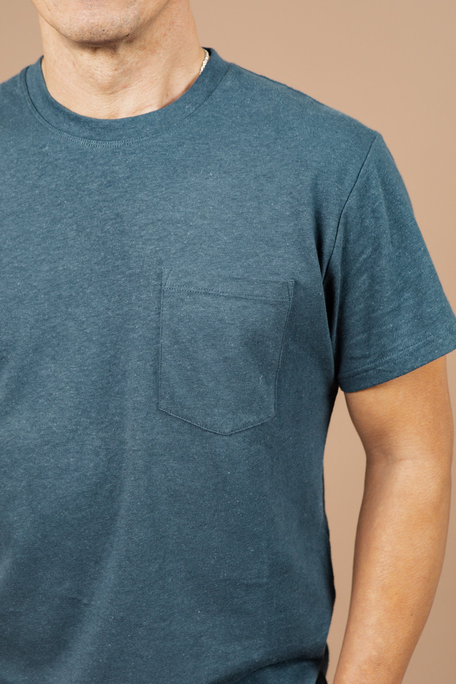 A person wears the Bridge & Burn Organic Hemp Pocket Tee in Storm Blue, featuring a chest pocket. Only their upper torso and part of their arms are visible against a plain beige background.