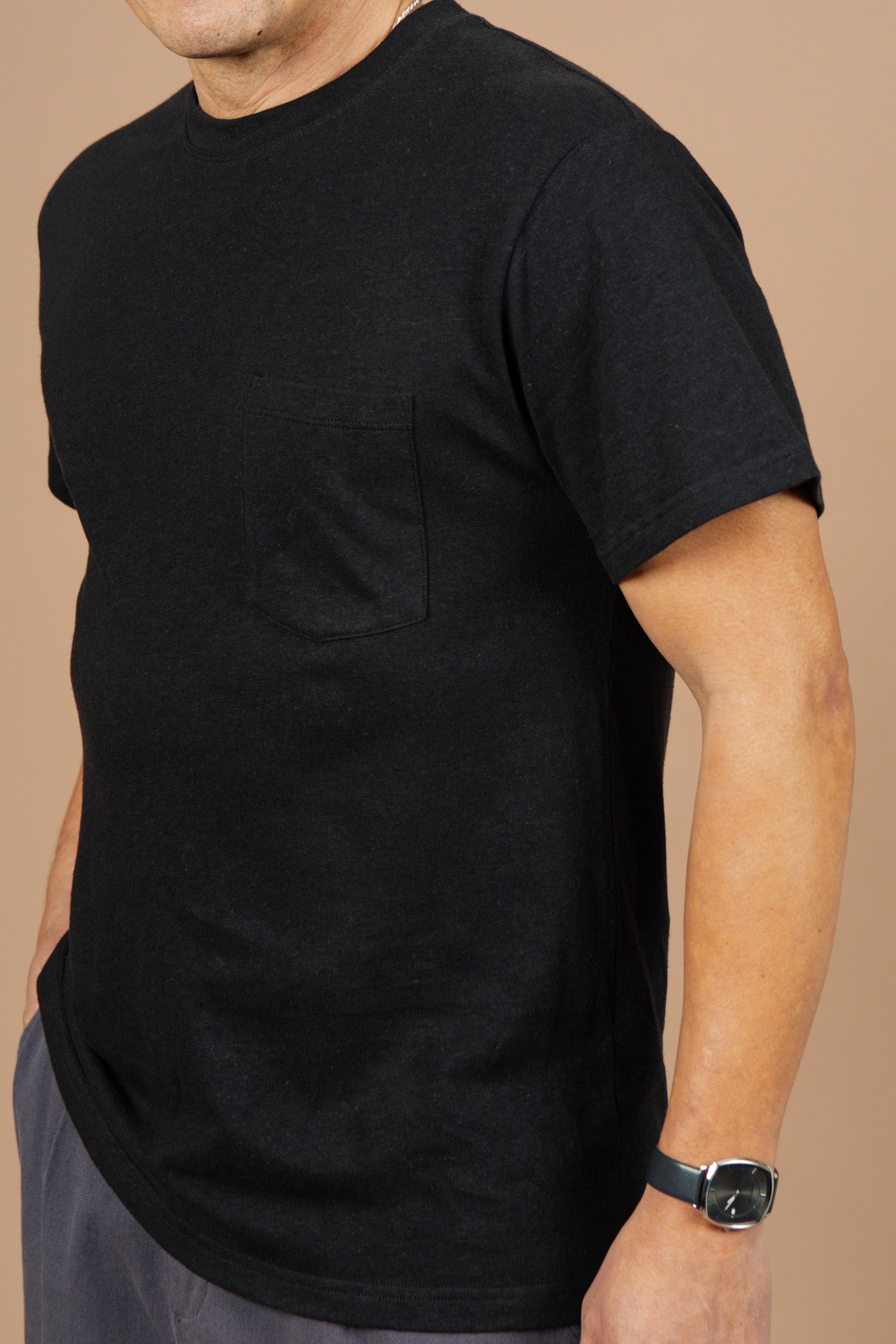 A person in a Bridge & Burn Organic Hemp Pocket Tee in Washed Black, gray pants, and a black wristwatch stands against a beige background with one hand in their pocket; their face is partially obscured.