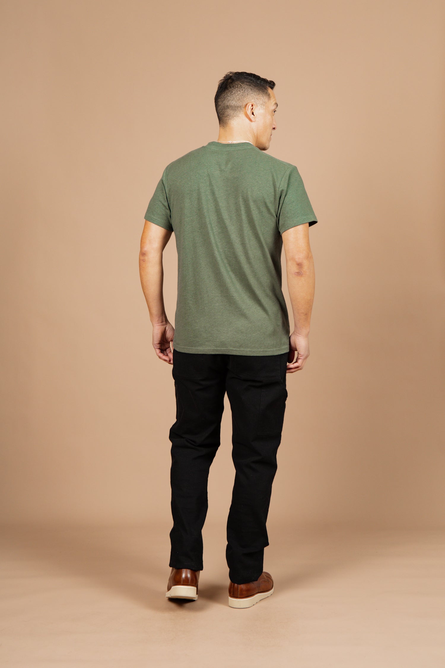 A man stands with his back to the camera against a beige background, wearing Bridge & Burn's Organic Hemp Pocket Tee in Cypress, along with black pants and brown shoes. His arms hang naturally at his sides.