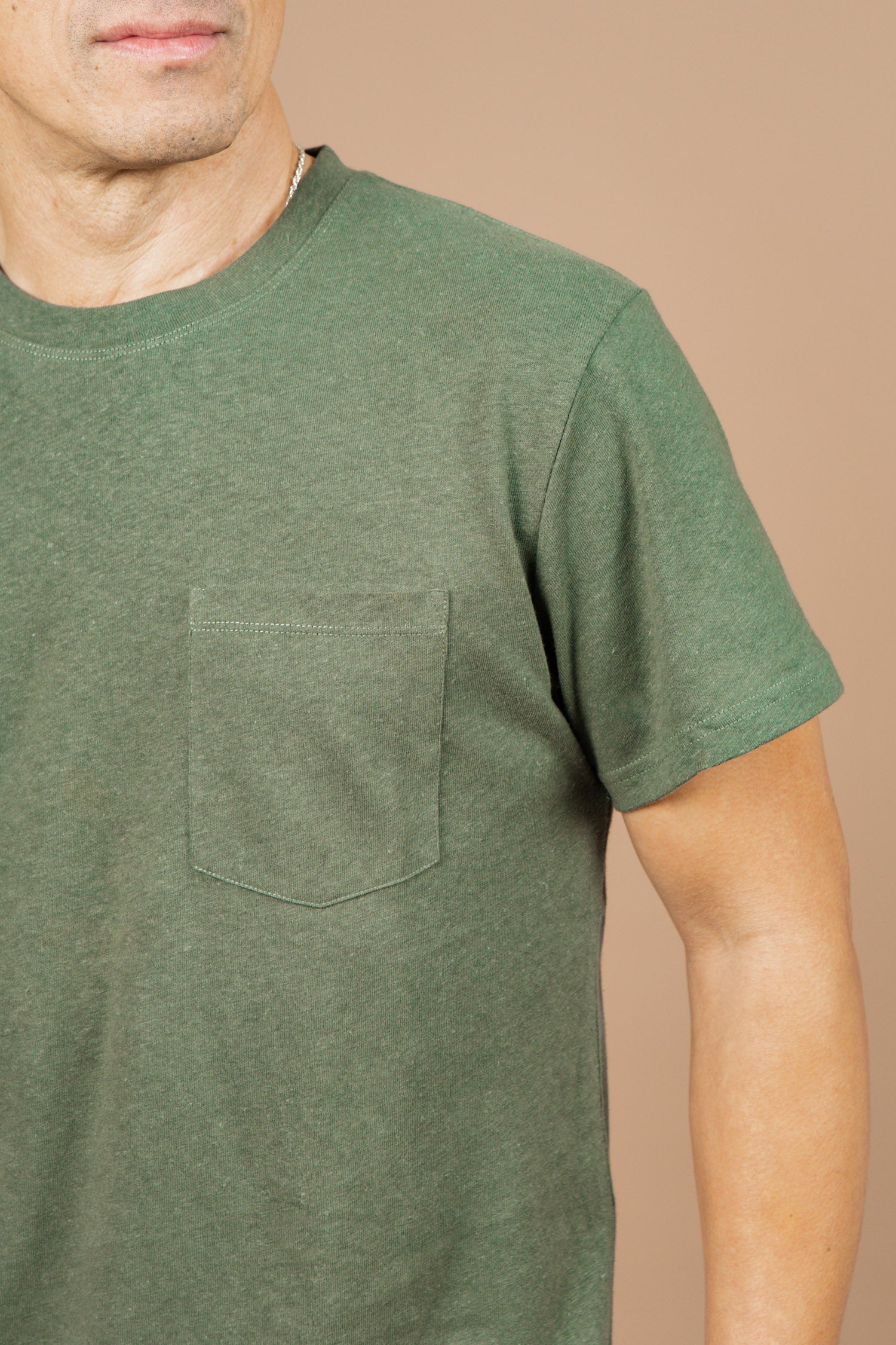 A man wears the Bridge & Burn Organic Hemp Pocket Tee in Cypress, featuring a chest pocket, standing against a beige background. Only his upper body and part of his face are visible.