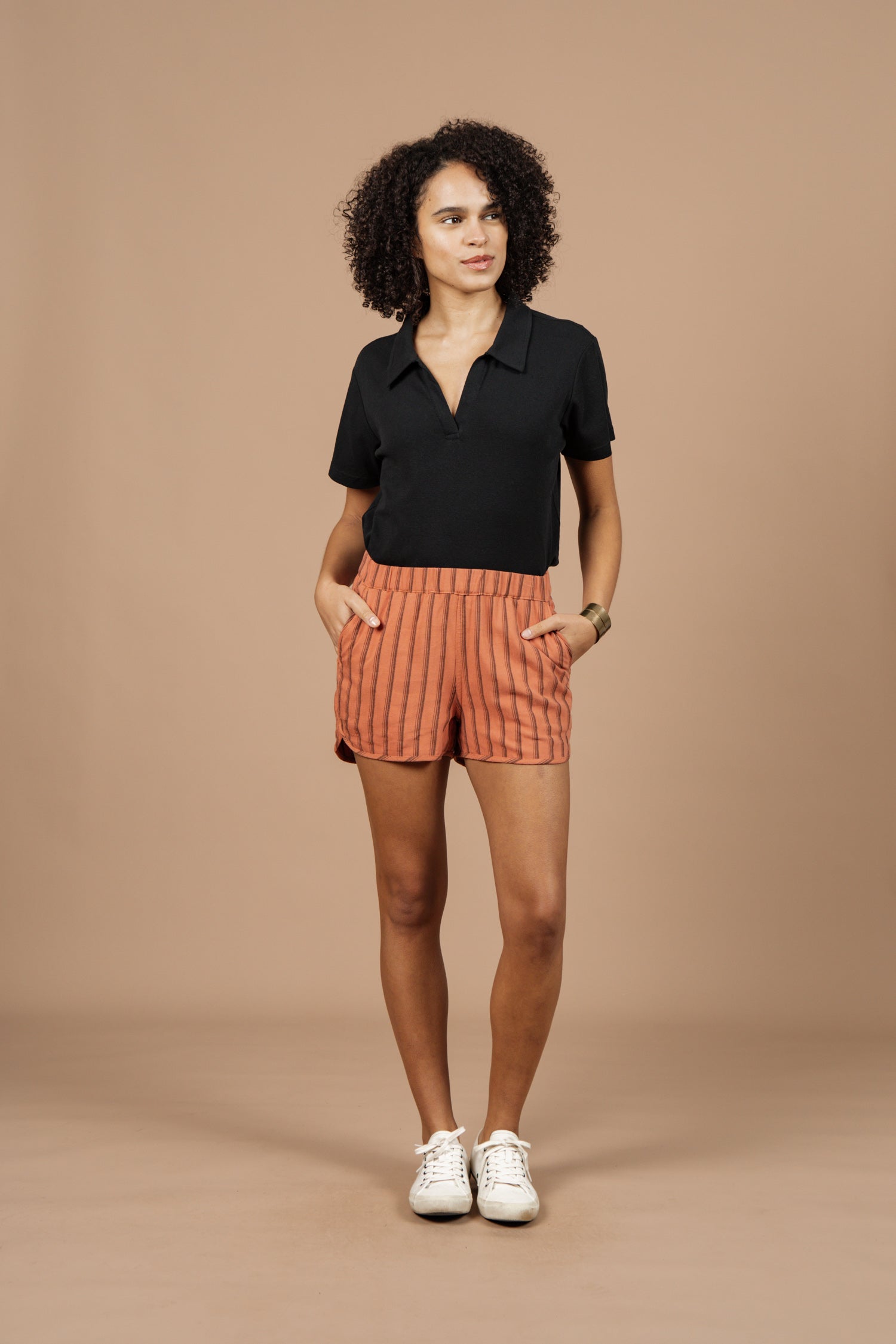 A woman stands against a tan background wearing Bridge & Burn's Luca Track Short in Rust Stripe, paired with a black short-sleeve polo, white sneakers, and a bracelet, with her hands in her pockets.