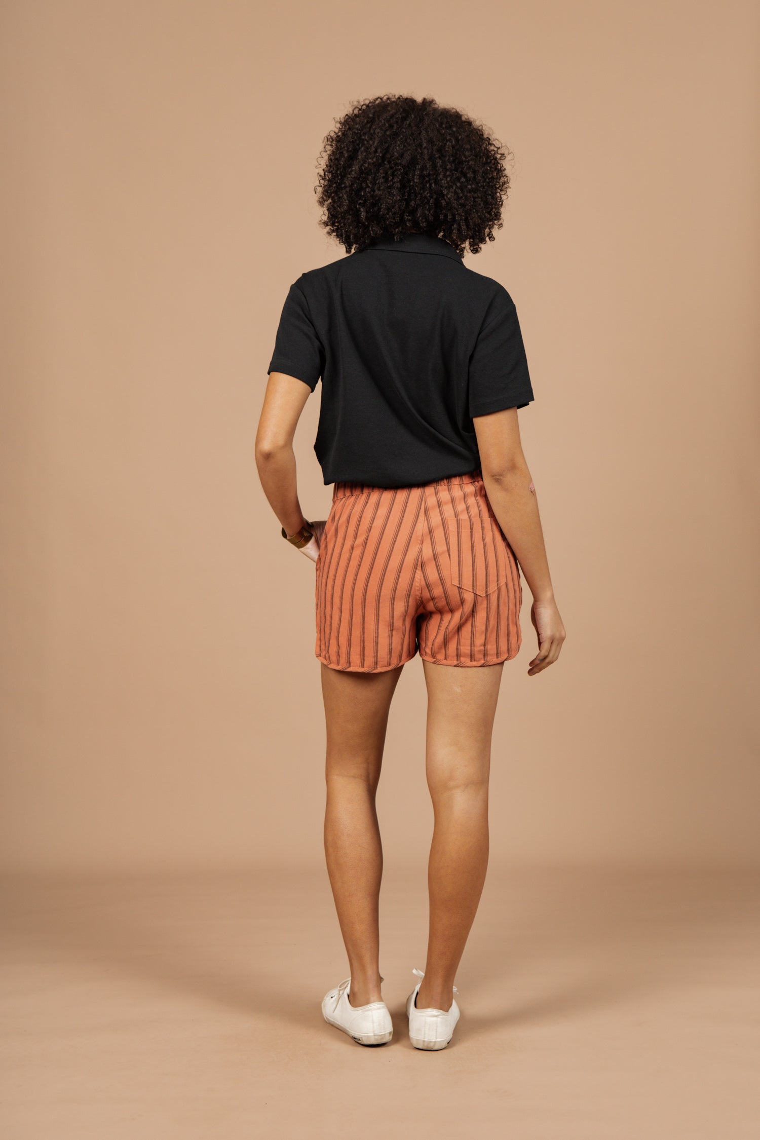 A person with curly hair, wearing a black shirt, Bridge & Burn Luca Track Short in Rust Stripe, and white shoes stands facing away from the camera against a plain beige background.