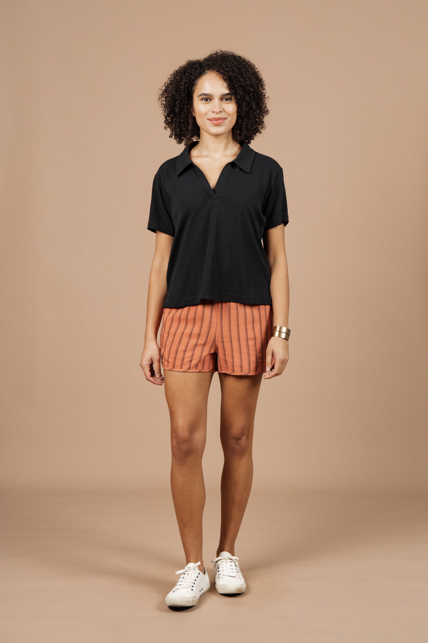 A person stands against a plain beige background, smiling with hands relaxed at their sides, wearing the Bridge & Burn Avery Boxy Top in black jersey, orange striped shorts, white sneakers, and a gold bracelet.