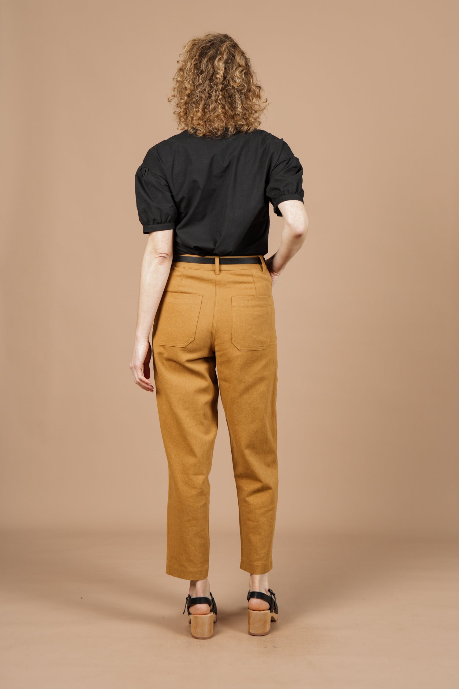 A person with curly hair, wearing the Bridge & Burn Charlotte Shirt in black poplin, tan high-waisted pants, and black sandals, stands facing away against a solid beige background.