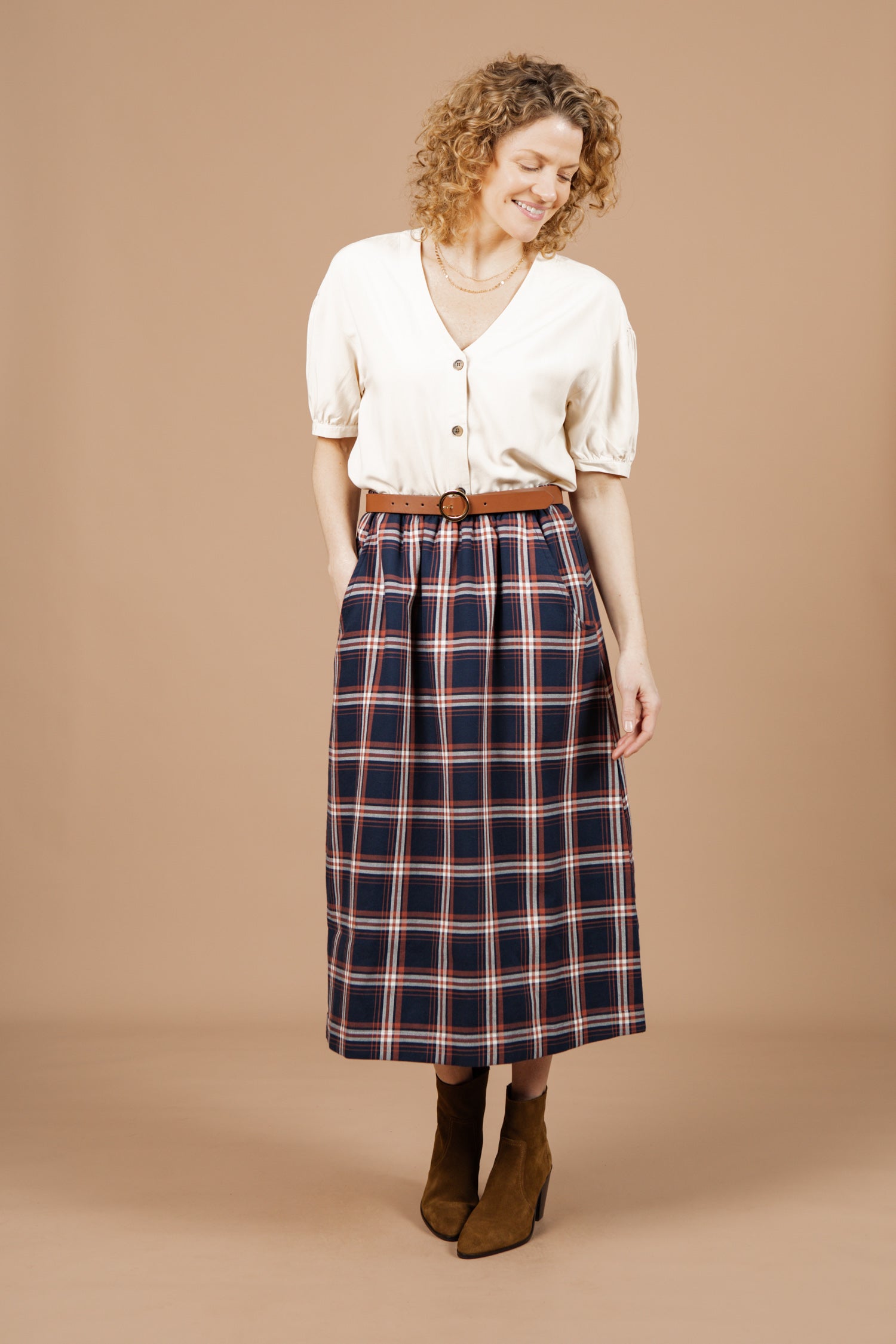 A woman with curly hair smiles against a beige background, wearing the Charlotte Shirt in cream poplin by Bridge & Burn, styled with a high-waisted navy plaid midi skirt and brown suede ankle boots.